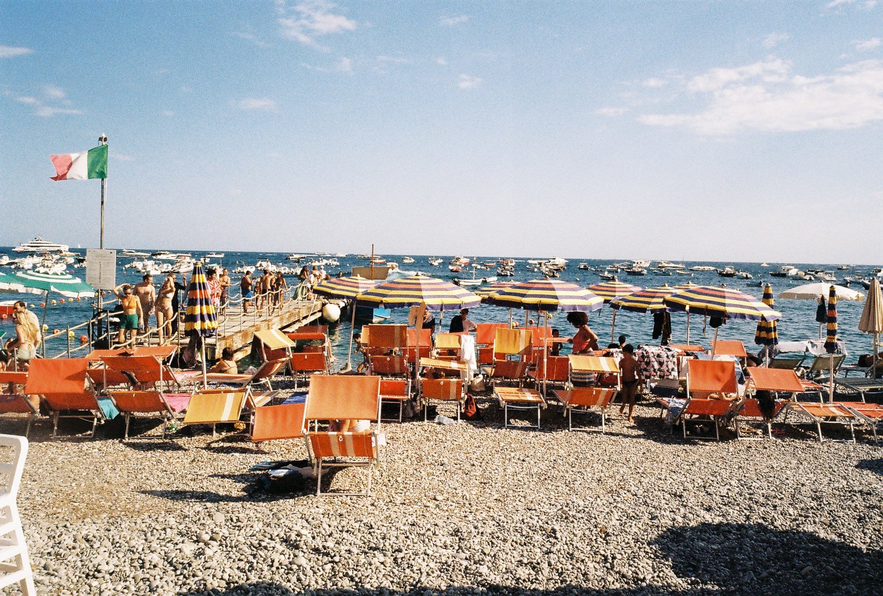 A crowded beach scene with orange lounge chairs and colorful umbrellas on pebbled shore, people swimming and walking on a pier, with numerous boats in the blue ocean and a partly cloudy sky.