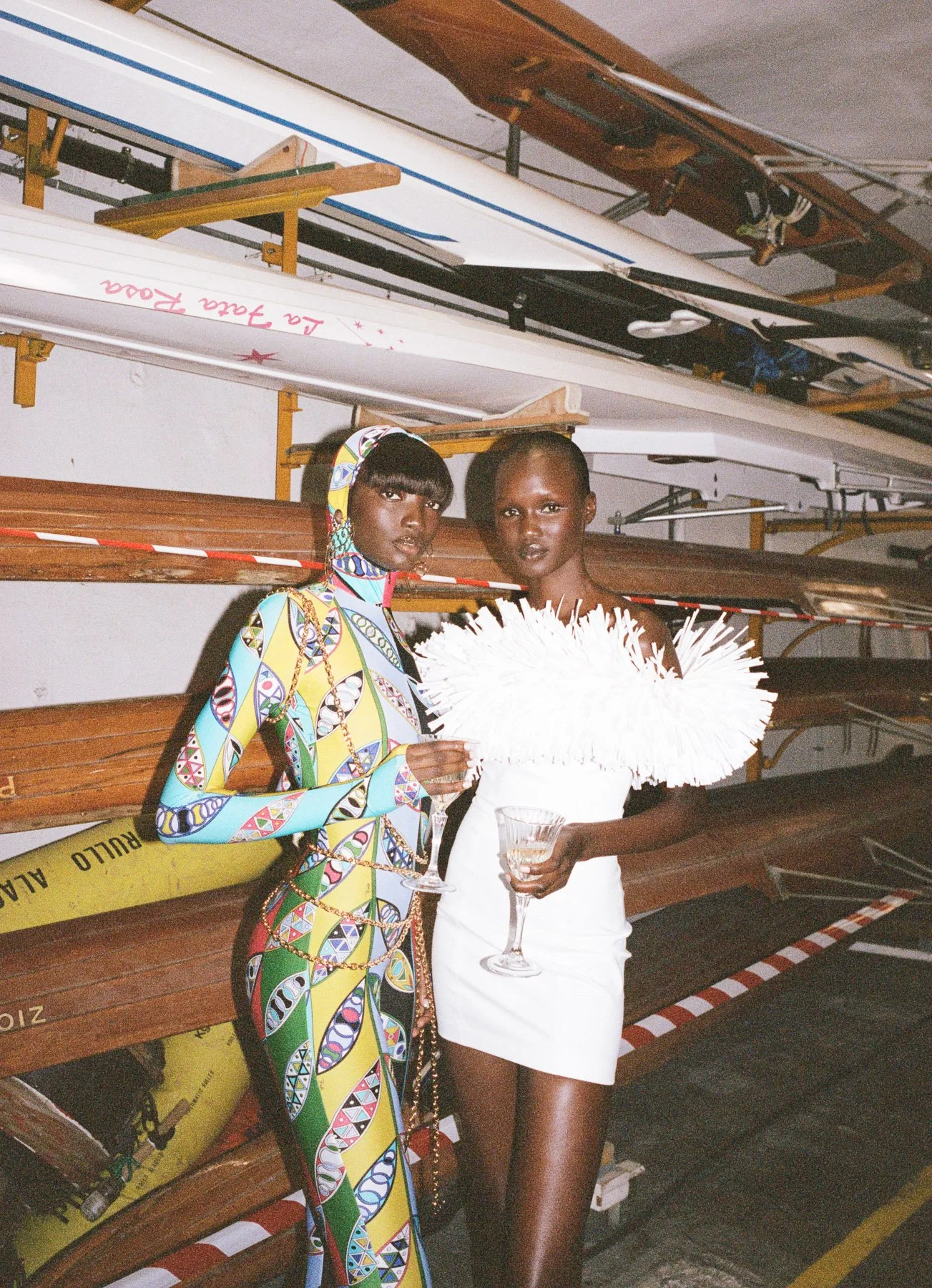 Two women standing in front of wooden canoes stacked on a rack. One woman wears a colorful patterned dress and headscarf, holding a wine glass. The other woman is dressed in a white outfit, holding a wine glass with a large white feathered accessory.