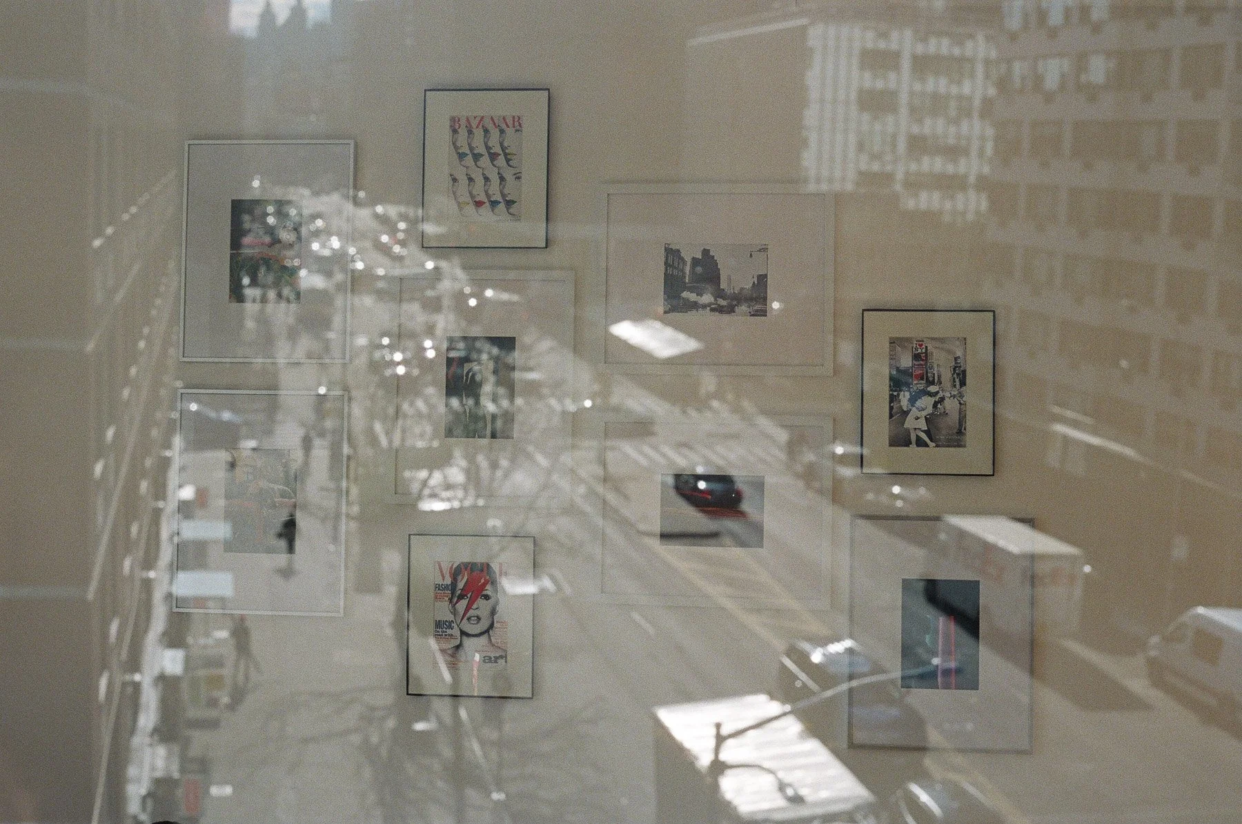 Reflection of urban street scene with framed artwork on wall, including magazine cover with stylized female face, and outdoor architecture visible through window.