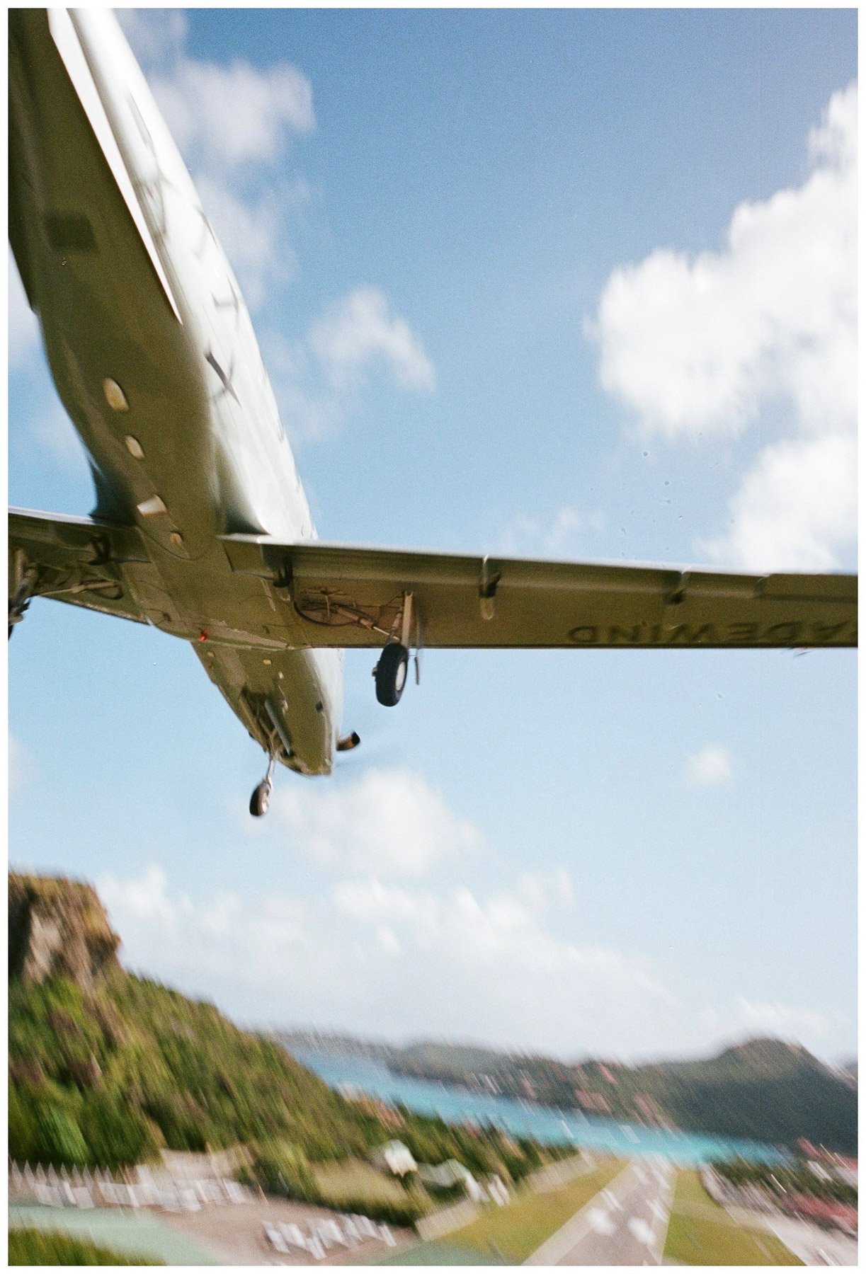 An airplane flying low over a coastal town with lush green hills, residential area, and coastline in the background under a partly cloudy sky.