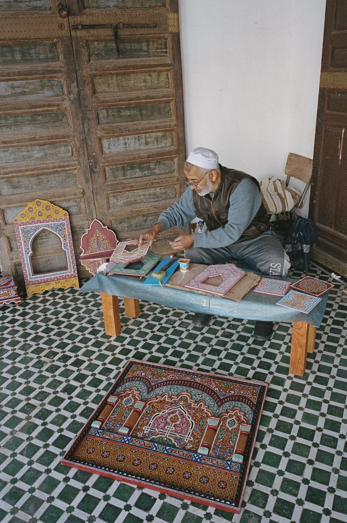 An elderly man wearing a white cap, glasses, and a vest sitting on a small chair, working on a craft project involving intricate, colorful painted wooden pieces at a low table covered with a plastic sheet. The setting appears to be a traditional room