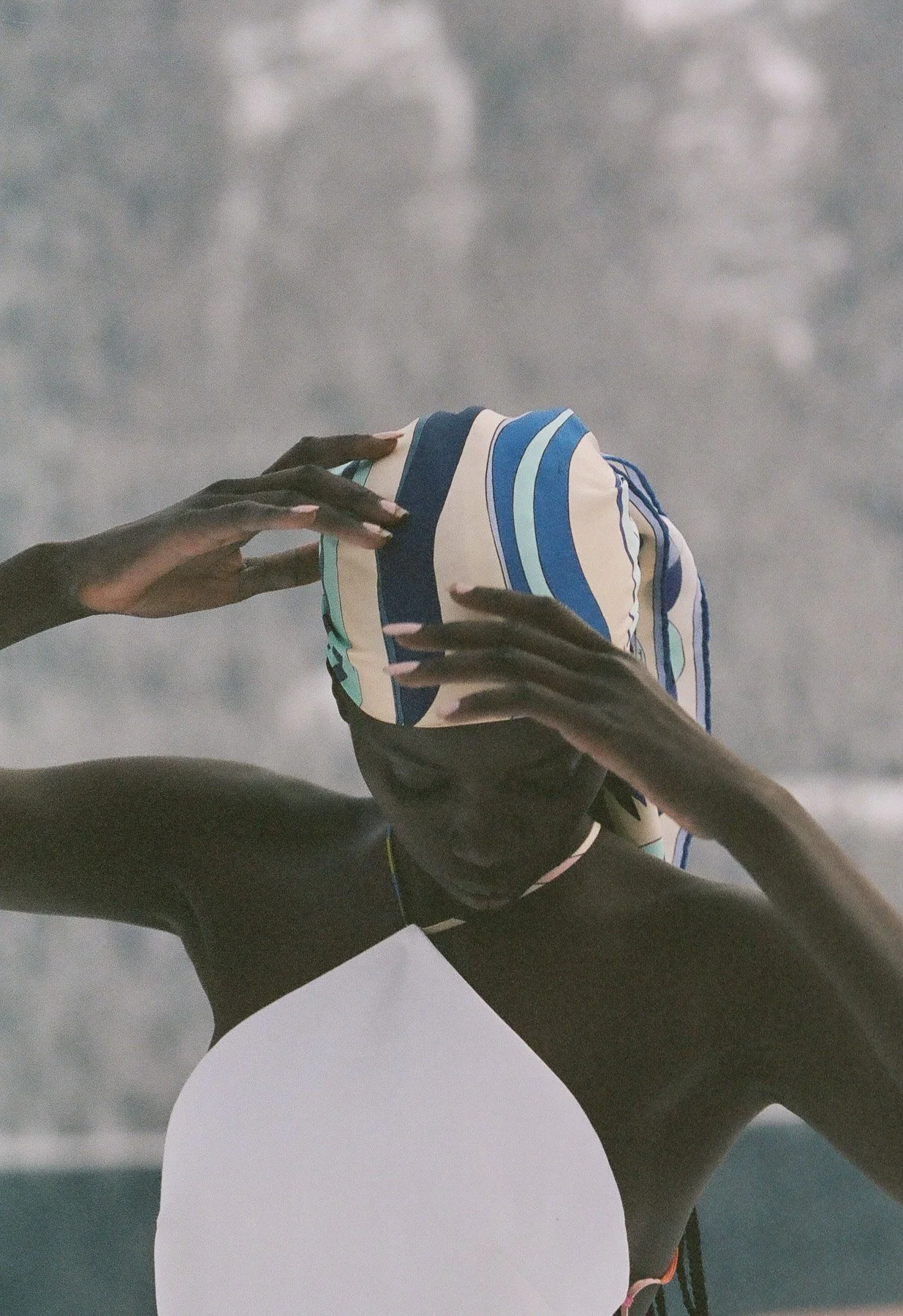 A person with dark skin pulling a striped fabric over their head at the beach, the ocean waves in the background, wearing a black swimsuit.