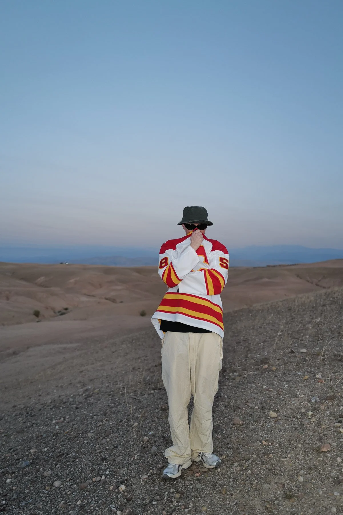 A person standing in a barren, rocky landscape under a clear blue sky. They are wearing a hockey jersey, beige pants, sneakers, a bucket hat, and sunglasses, with their face partially covered by their hand.