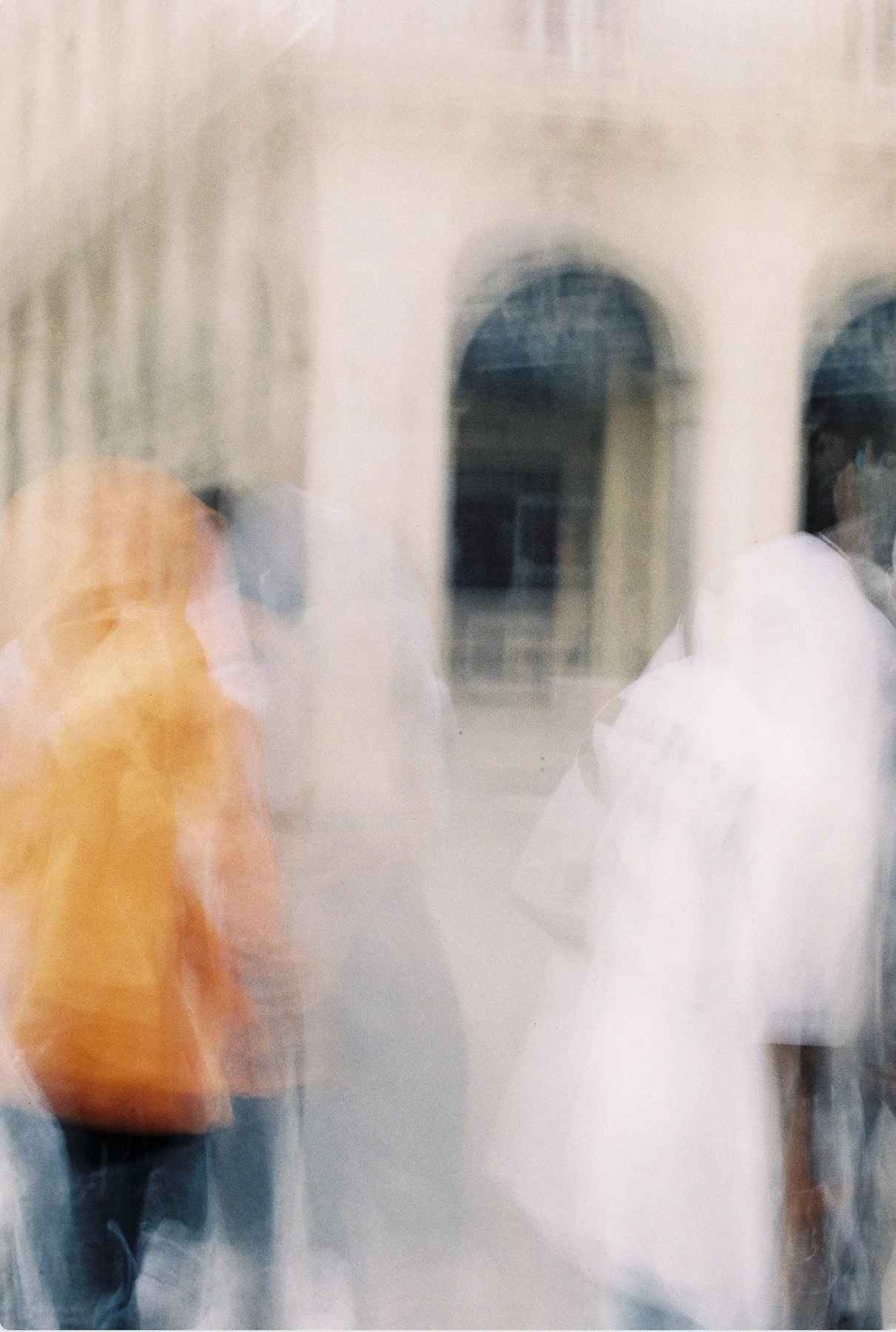 Blurry photo of people walking past a building with arches, seen through a glass window.