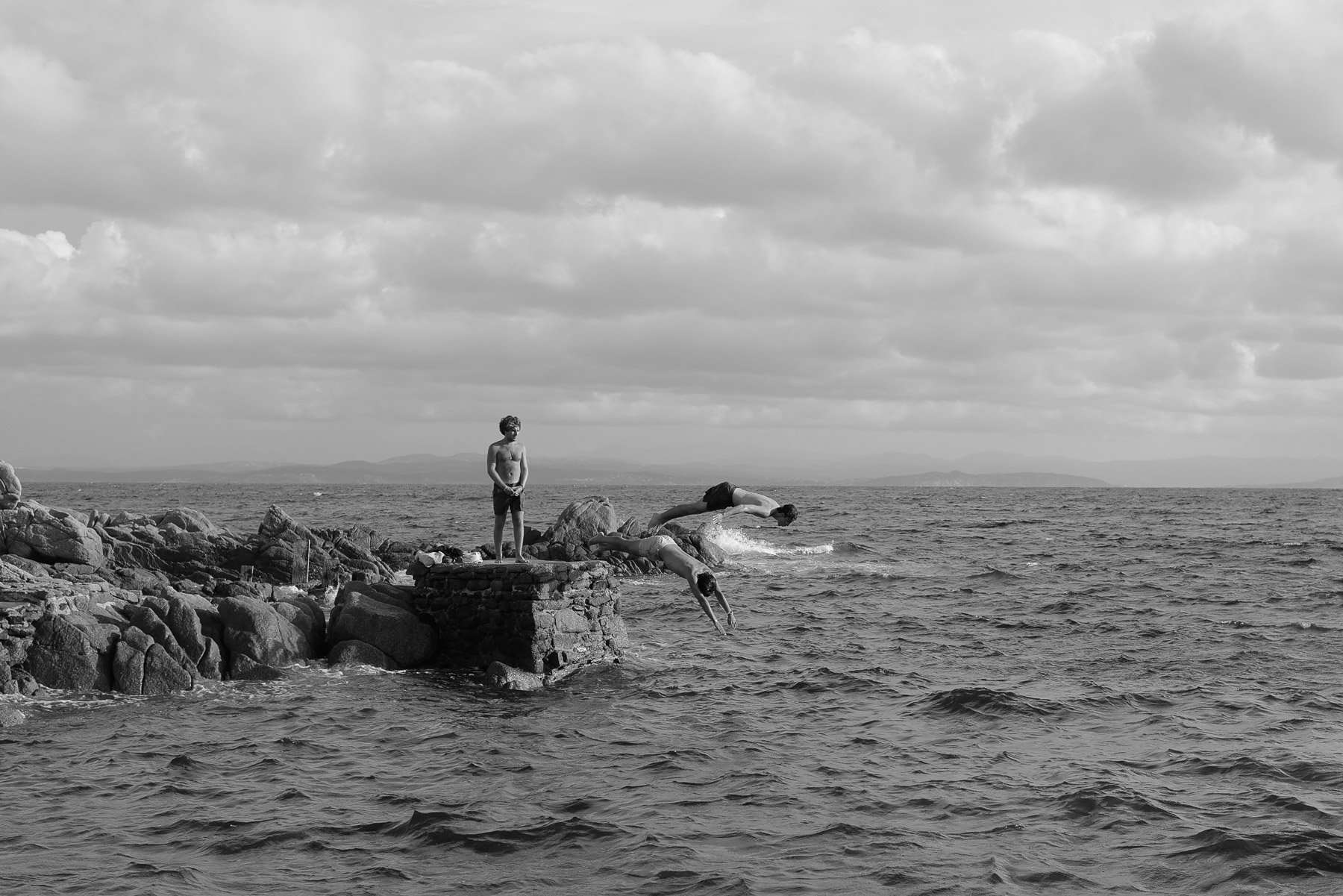 Four children in swimsuits diving off and standing on a rocky pier into the sea, with cloudy skies above and mountains in the distance.