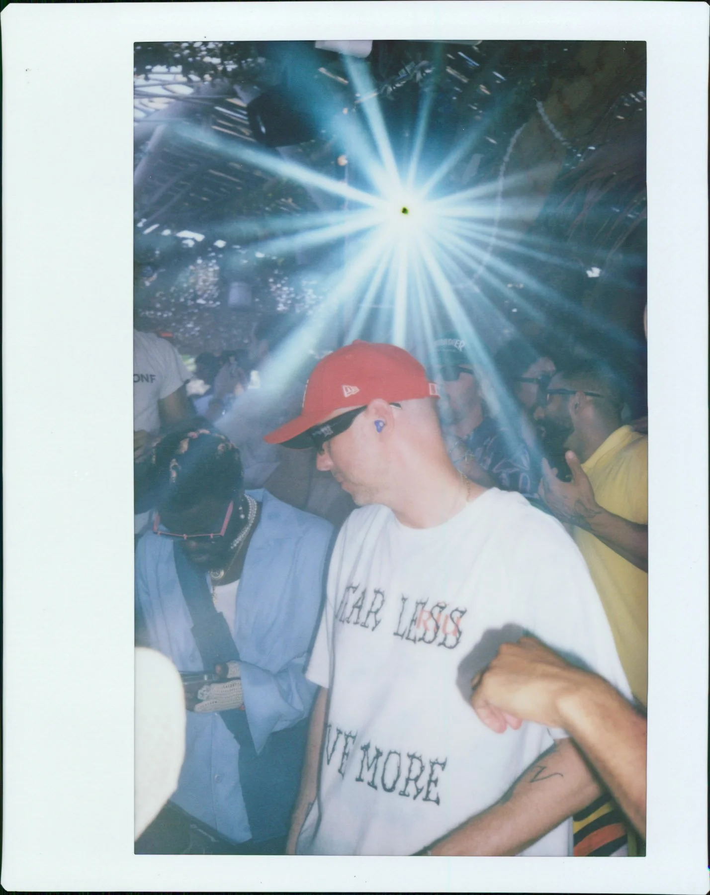 A group of people in a crowded indoor space with a bright light or laser beam shining from the ceiling, some wearing hats and sunglasses, and a person in the foreground with a tattooed arm and a white T-shirt.