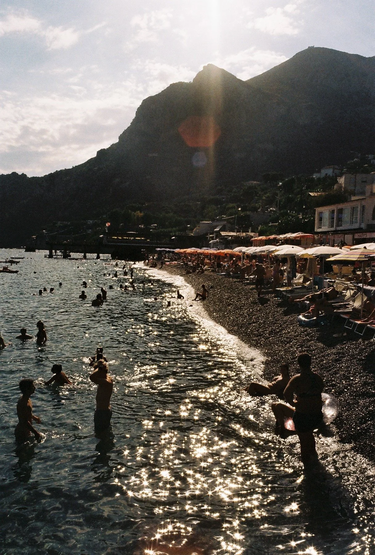 Beach with people swimming and lounging under umbrellas, mountain in the background, sun shining, creating sparkling reflections on the water.