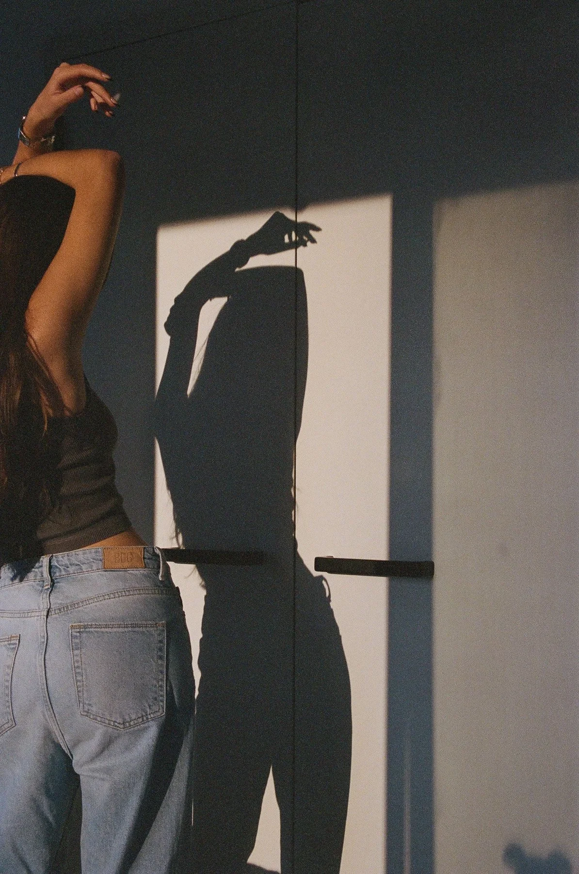 A woman wearing a black top and light blue jeans stands beside a mirror, creating a shadow of her hand on the wall.