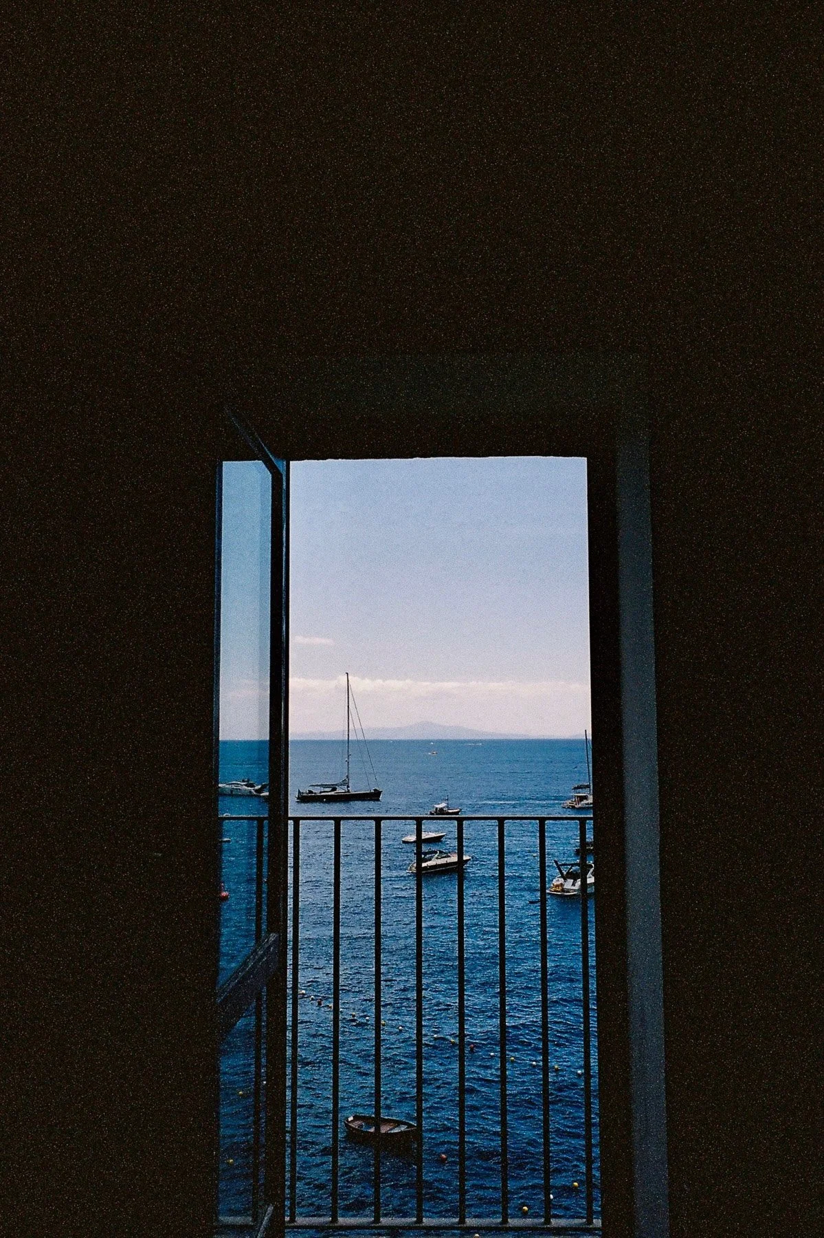 View of sailboats and the ocean through an open window with a balcony railing, under a clear blue sky.