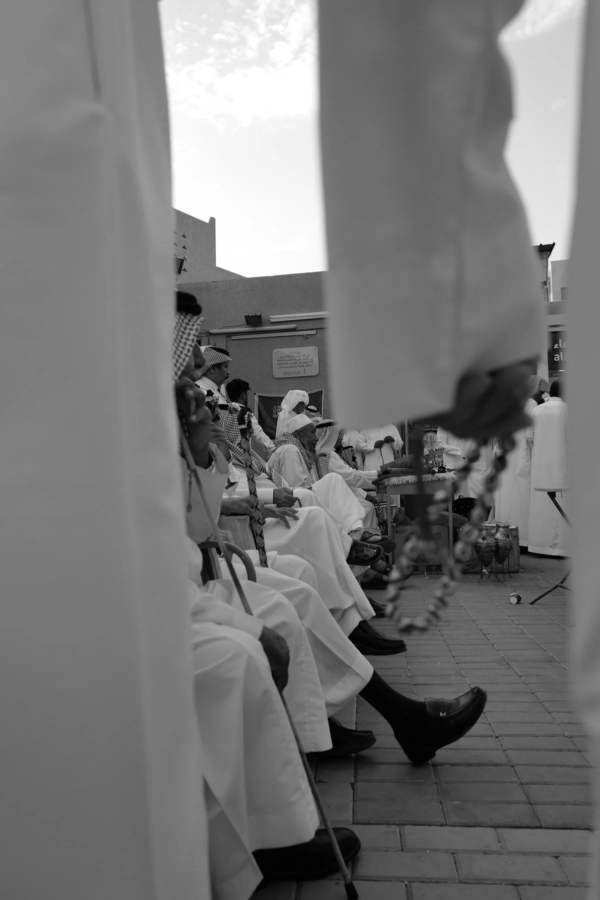 Black and white photo of a group of elderly men dressed in traditional white robes and head coverings, sitting outdoors, possibly participating in a ceremonial or cultural gathering. The photo is taken from a low angle through a narrow frame, with so