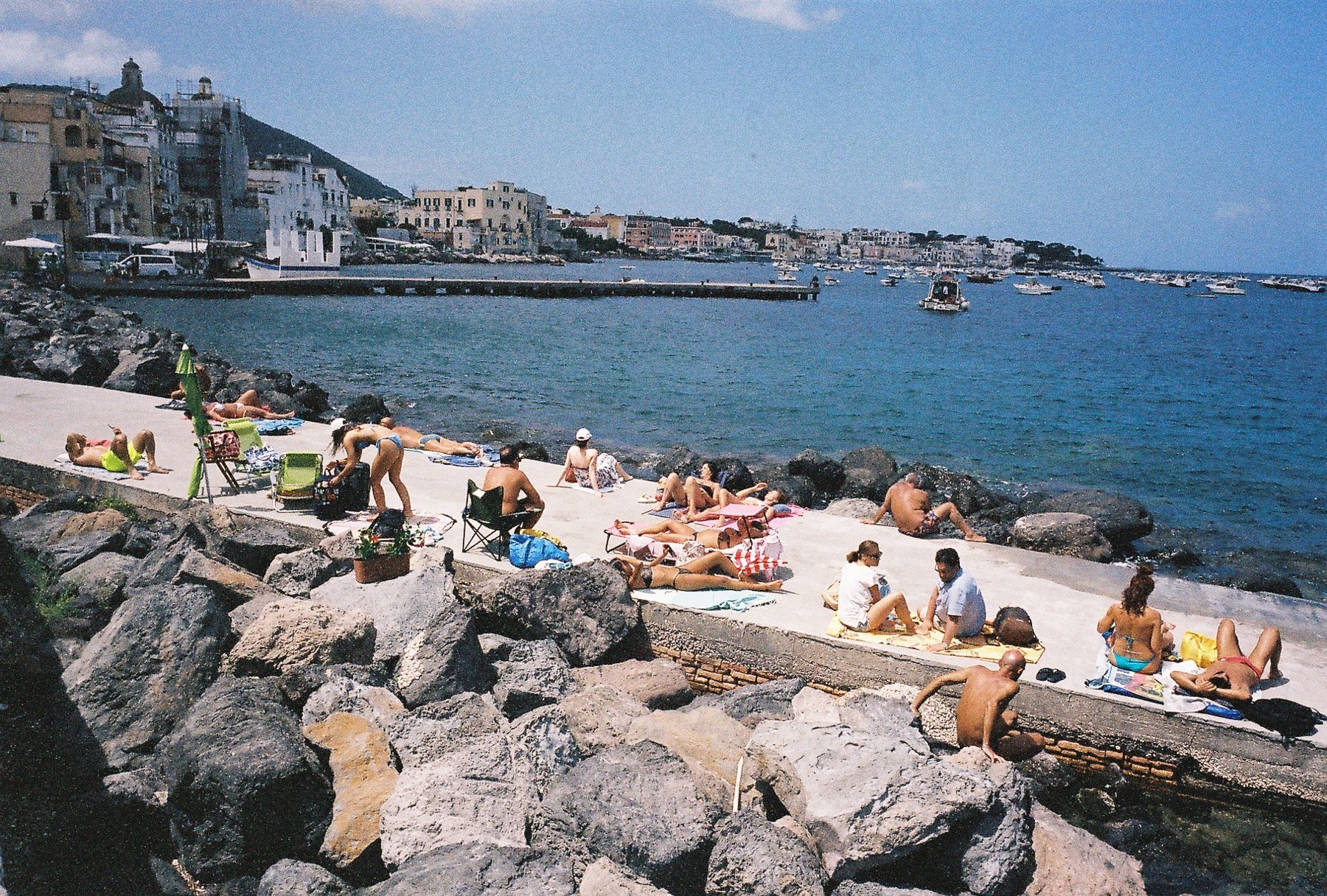 People relaxing and sunbathing on a seaside promenade with rocks and boats in the water, and buildings and hills in the background.