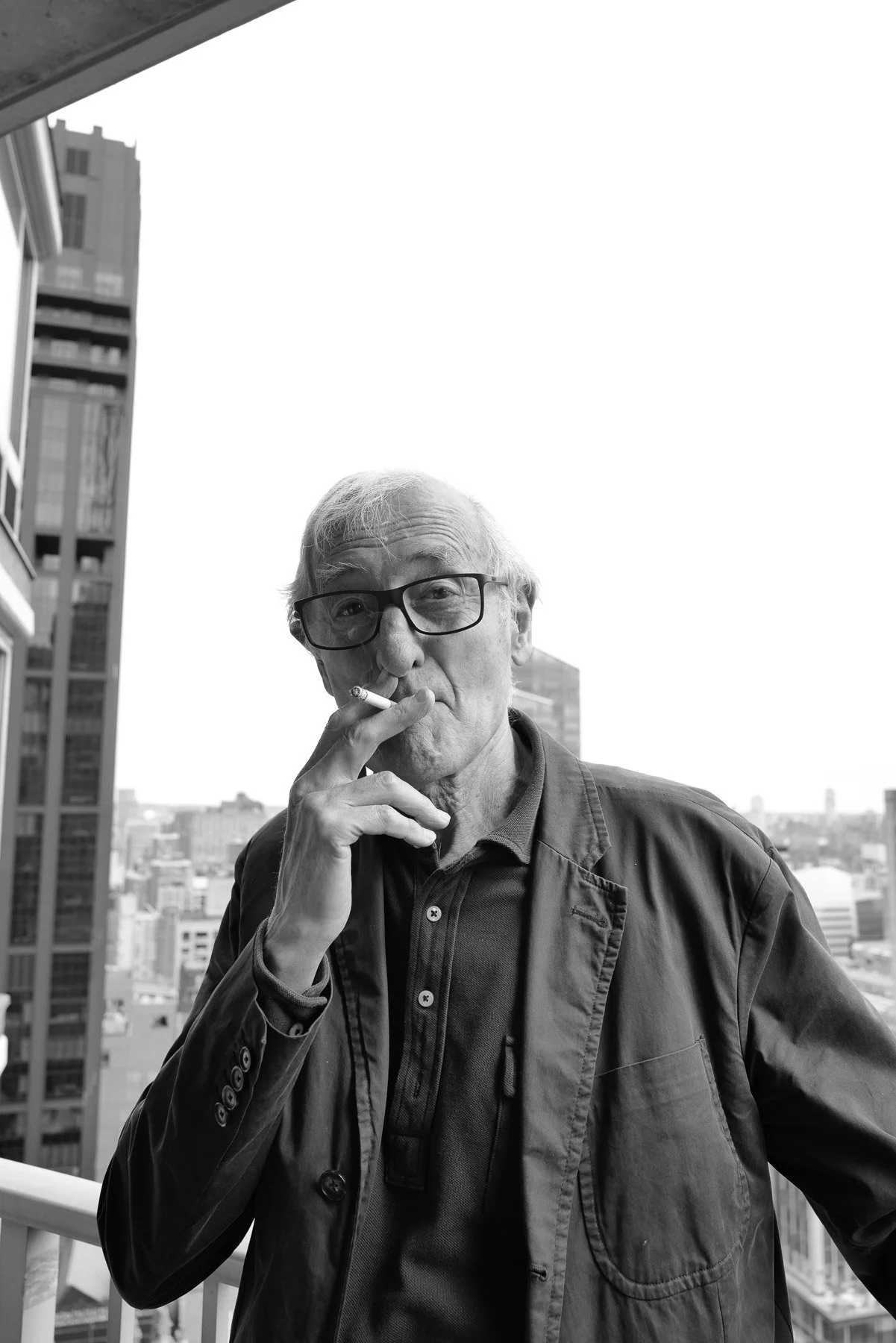 An elderly man with glasses smoking a cigarette on a balcony in an urban setting, with tall buildings in the background.