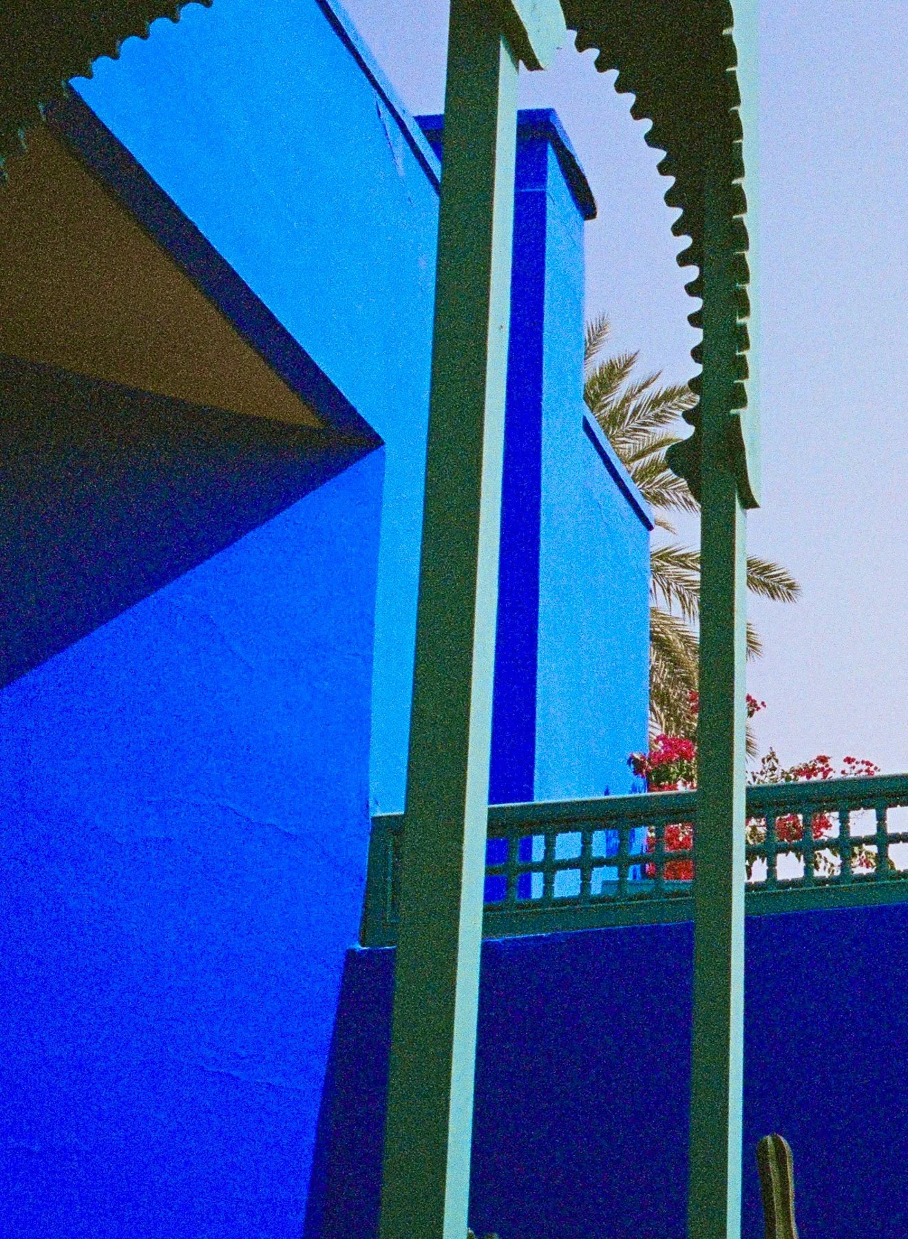 A close-up view of a vibrant blue building wall with green beams and a green fence, with palm trees and pink flowers in the background under a clear sky.