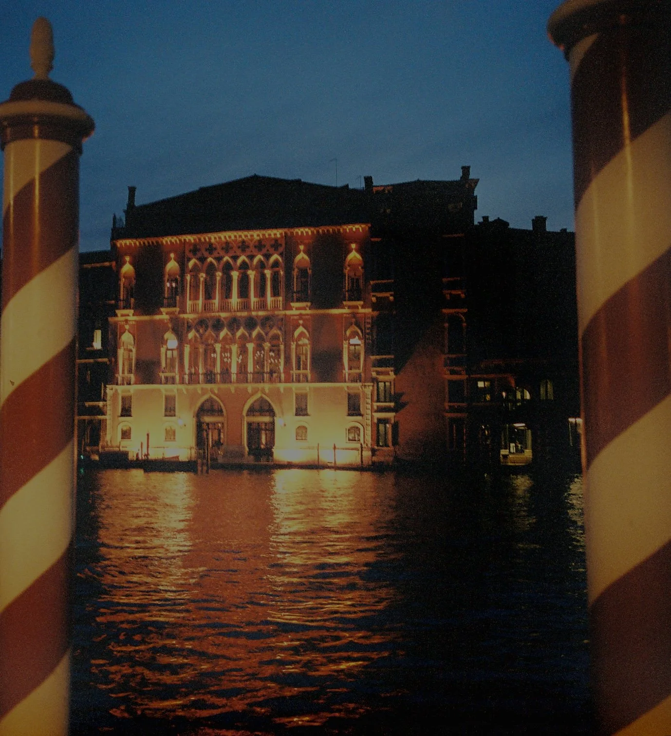 A nighttime scene of a historic building illuminated with warm lights, reflection on water in front, framed by striped red and white posts.