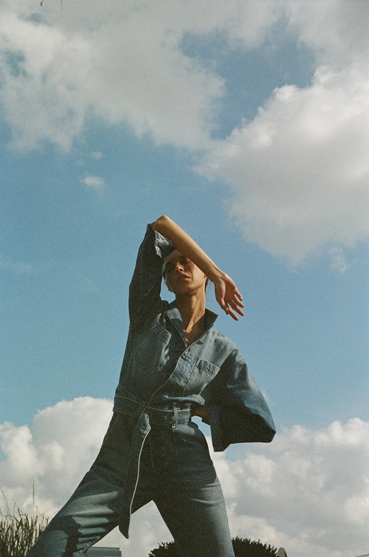 A person standing outdoors against a cloudy sky, wearing a denim jacket and black pants, with one arm raised over their face.