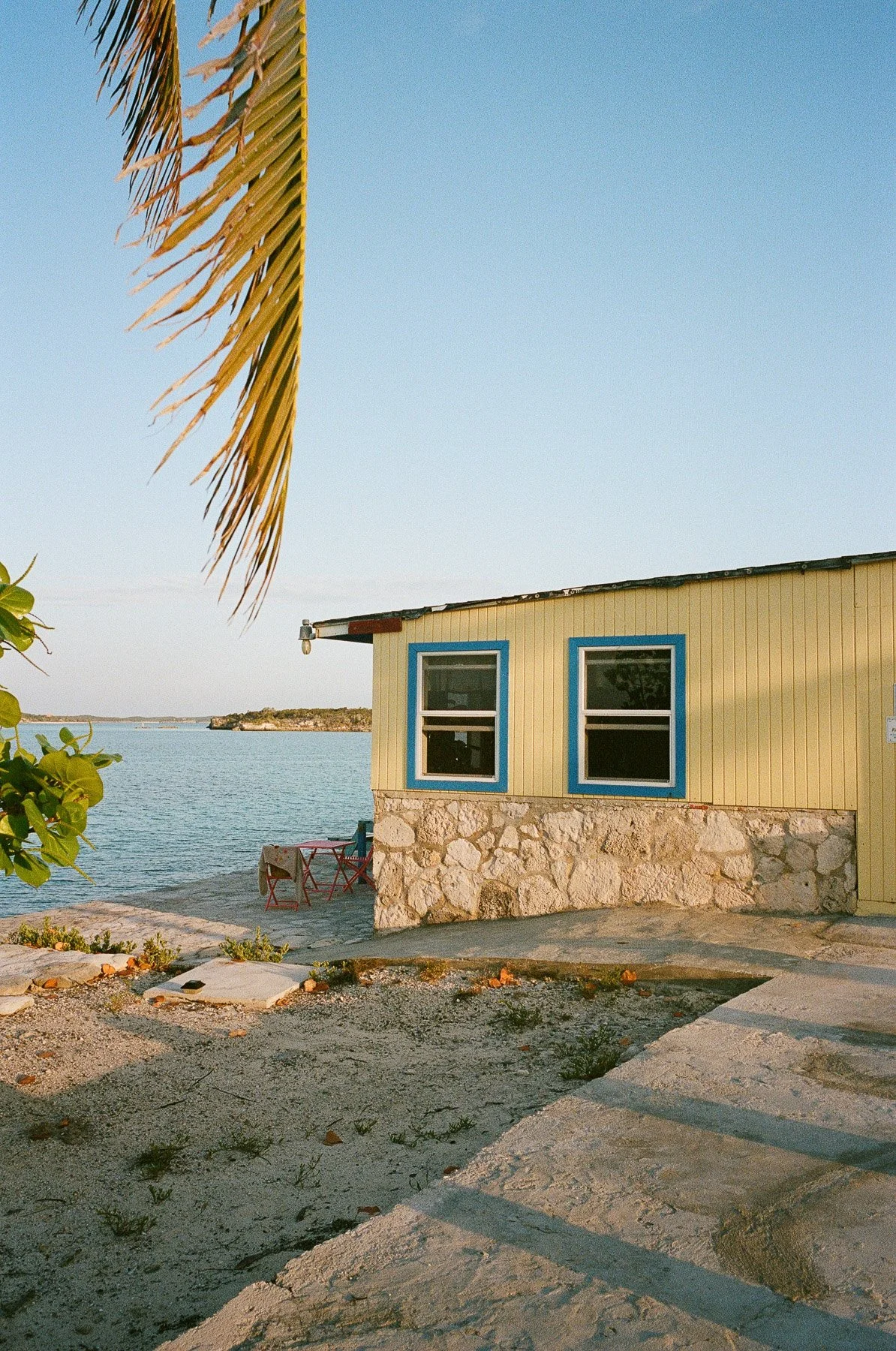 A yellow seaside house with blue-trimmed windows, made of stone and wood, next to calm water, with a small outdoor seating area under the sunlight, and a palm tree branch partially visible at the top left corner.