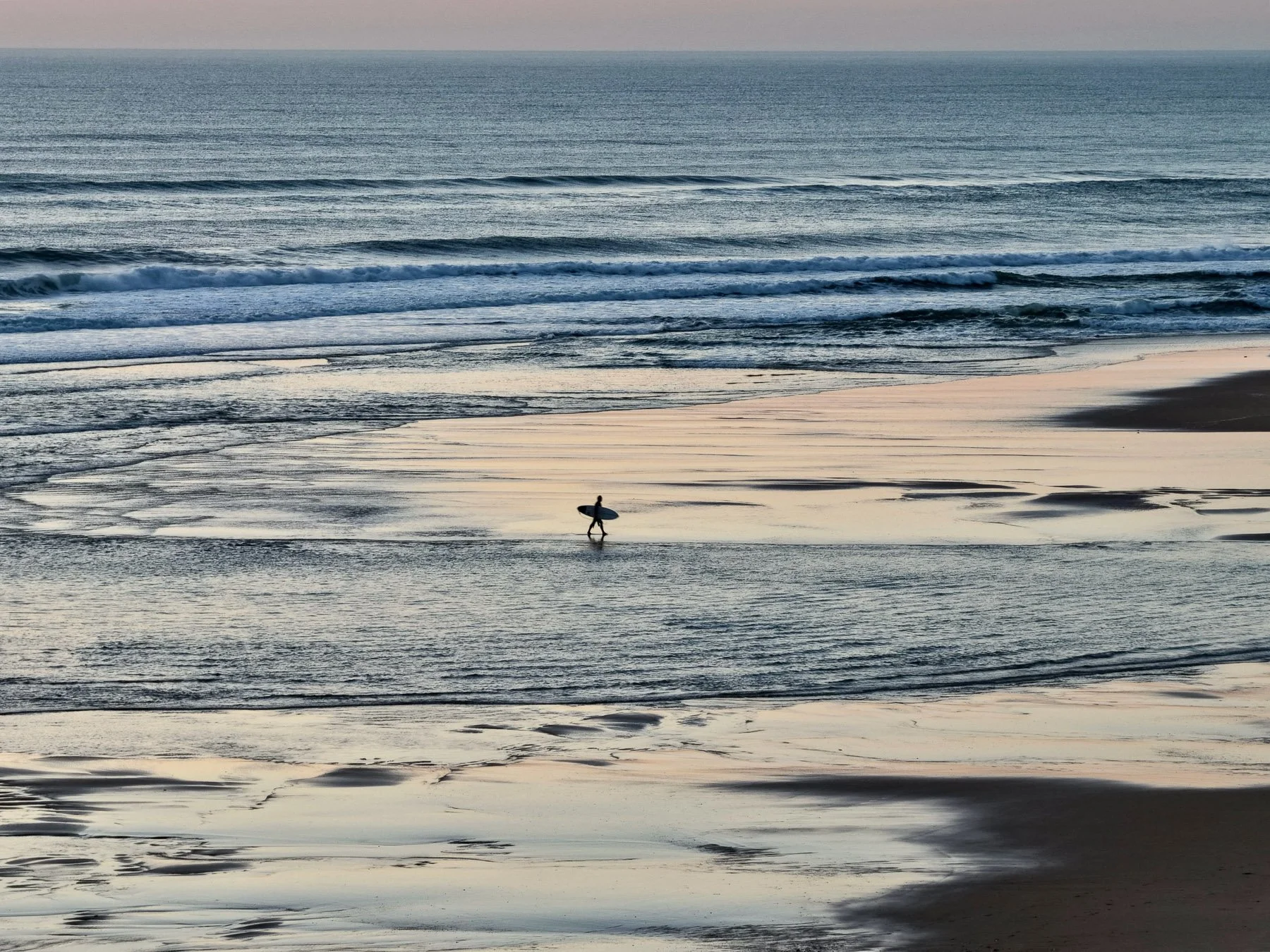 A surfer walking on the wet sand at a beach during sunset, carrying a surfboard under their arm, with gentle waves in the ocean in the background.