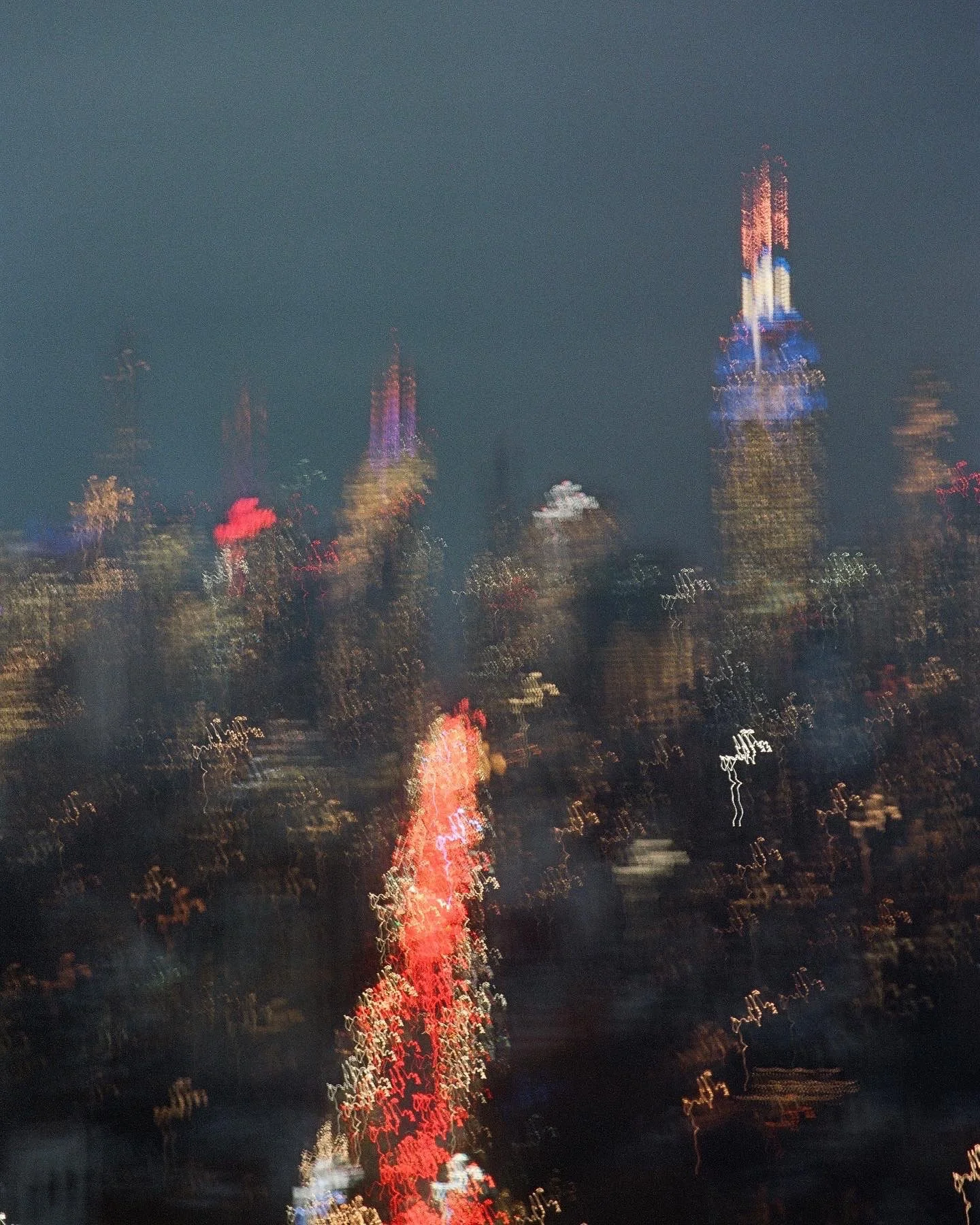 Night cityscape with blurred, colorful lights and tall buildings, including a prominent skyscraper with illuminated spires and a vertical white light.