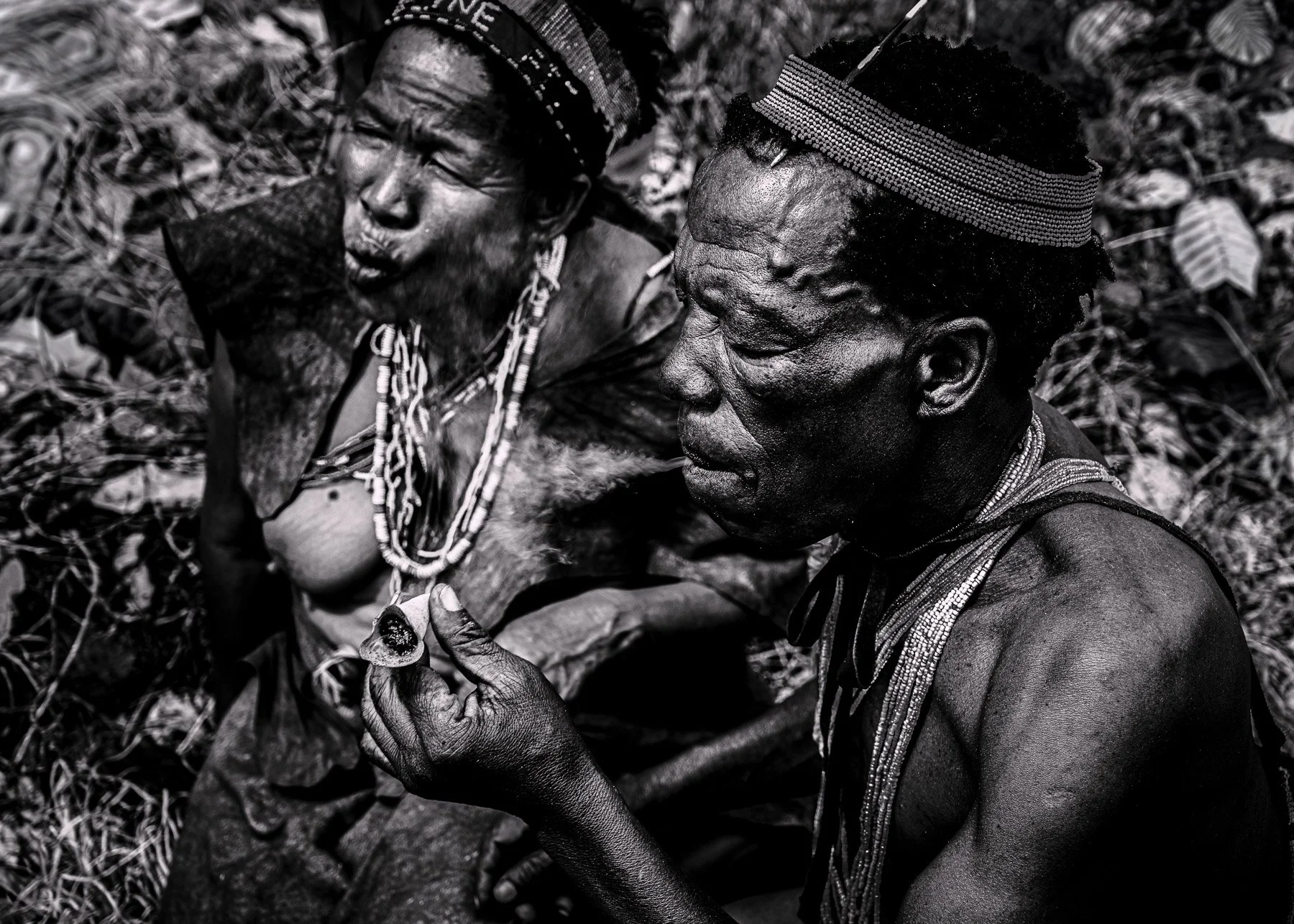 Two indigenous people outdoors, with one holding a small object and wearing traditional beaded jewelry and headdresses.