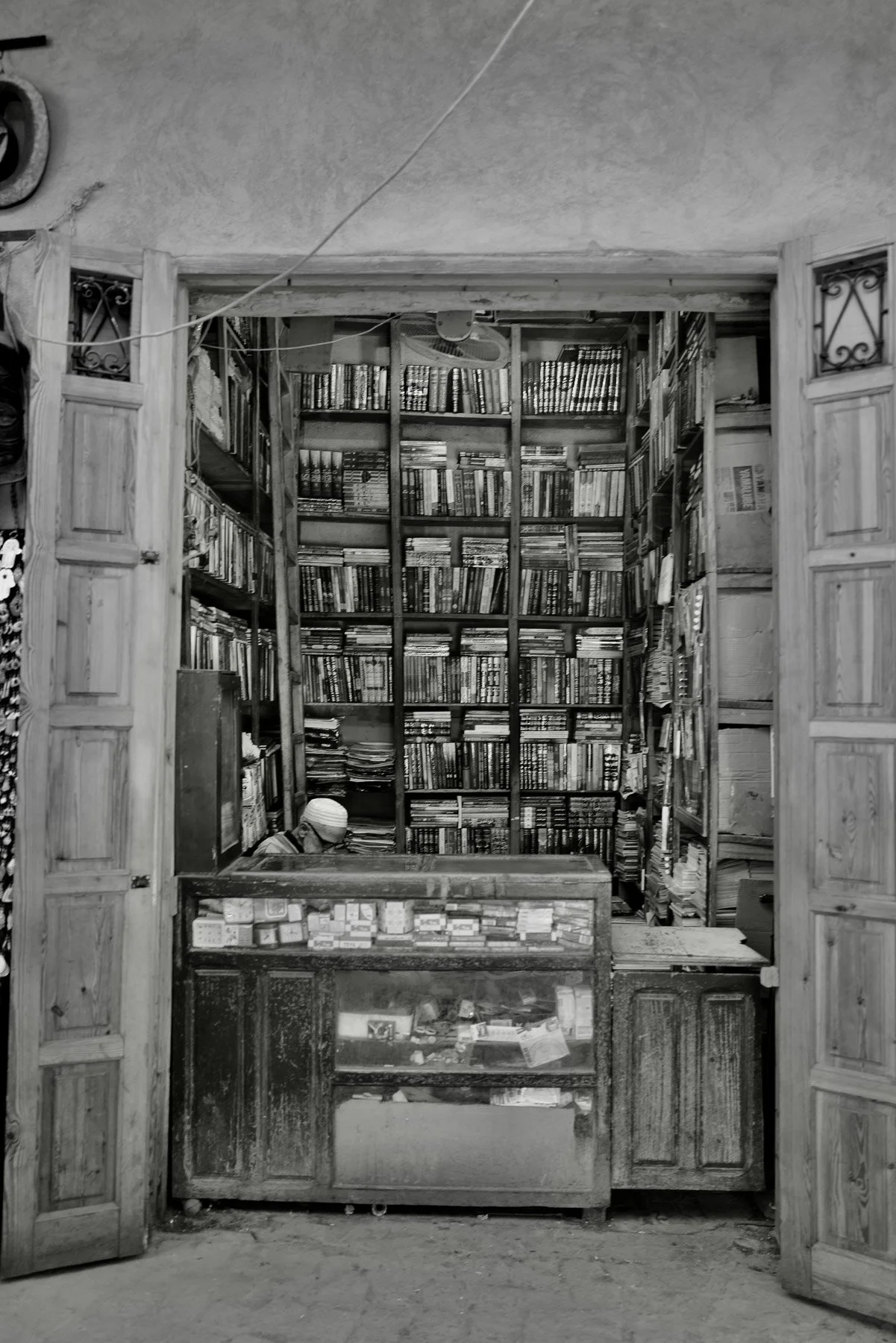 A small bookstore with wooden shelves filled with books. A person wearing a white cap is sitting behind the counter, which has glass display cases with items inside. The entrance is framed by open wooden doors with decorative metalwork. There are han