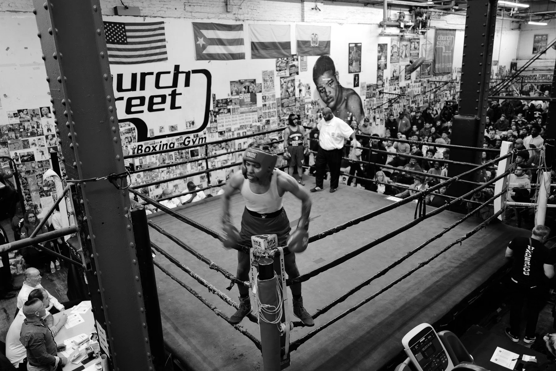 A boxing gym with an audience watching a boxer in the corner of the ring, wearing a helmet and shorts. The gym has American and Puerto Rican flags on the wall, and a large mural of a woman's face. Several people are seated or standing around the ring
