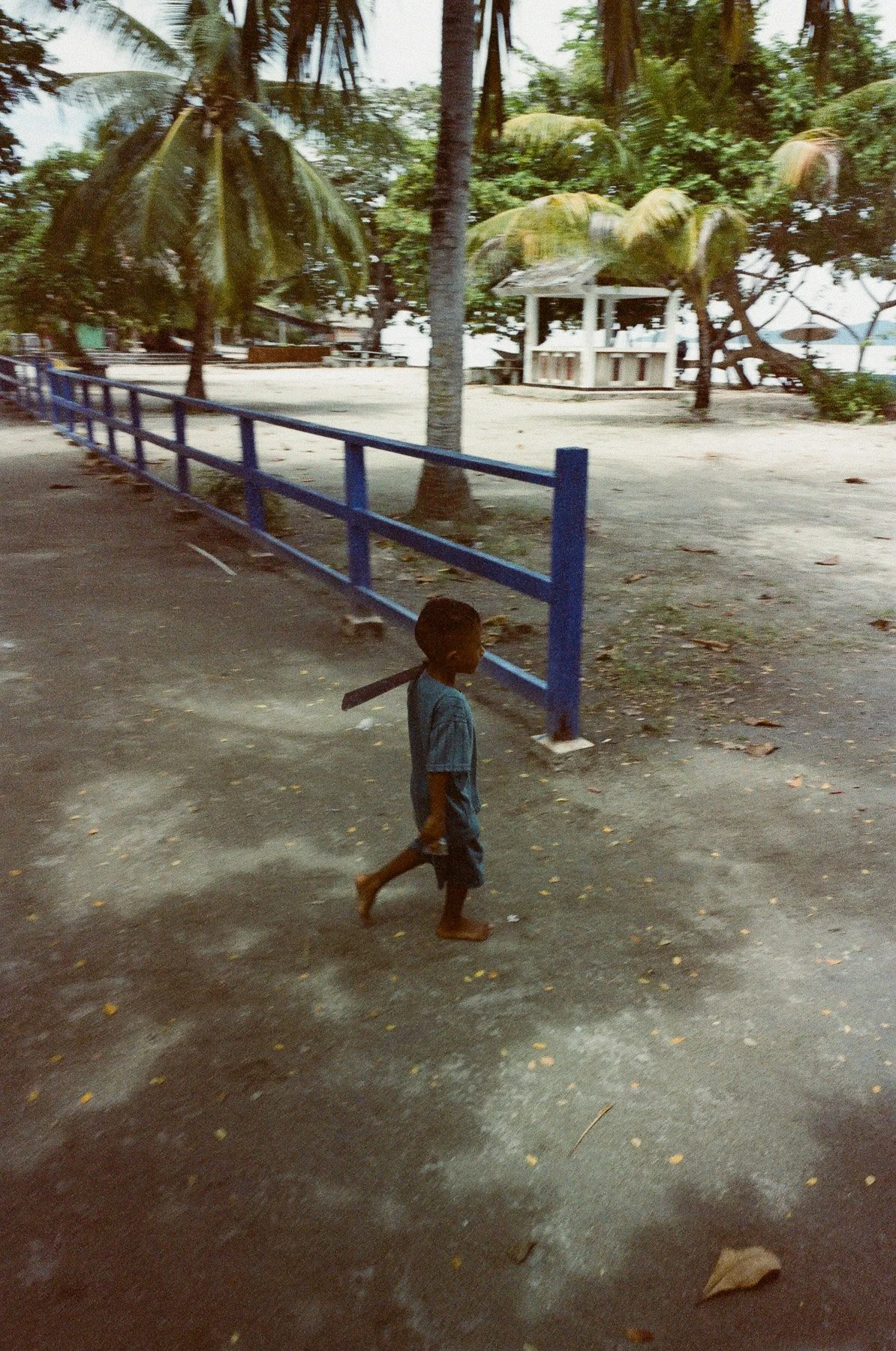 A young boy walking barefoot on a dirt path next to a blue metal fence, with trees and a small white structure in the background.