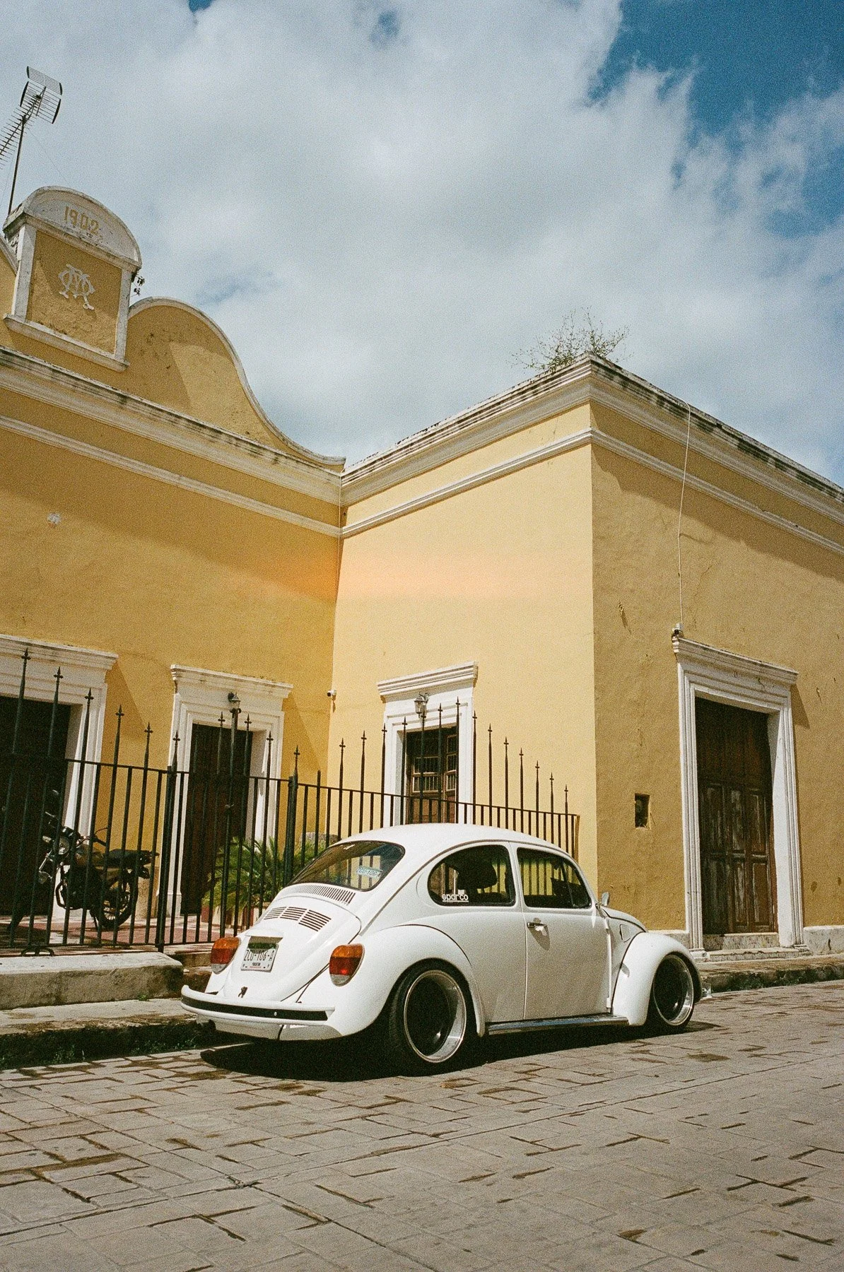 A vintage white Volkswagen Beetle parked on a cobblestone street in front of a yellow colonial-style building with white trim and barred windows, under a partly cloudy sky.