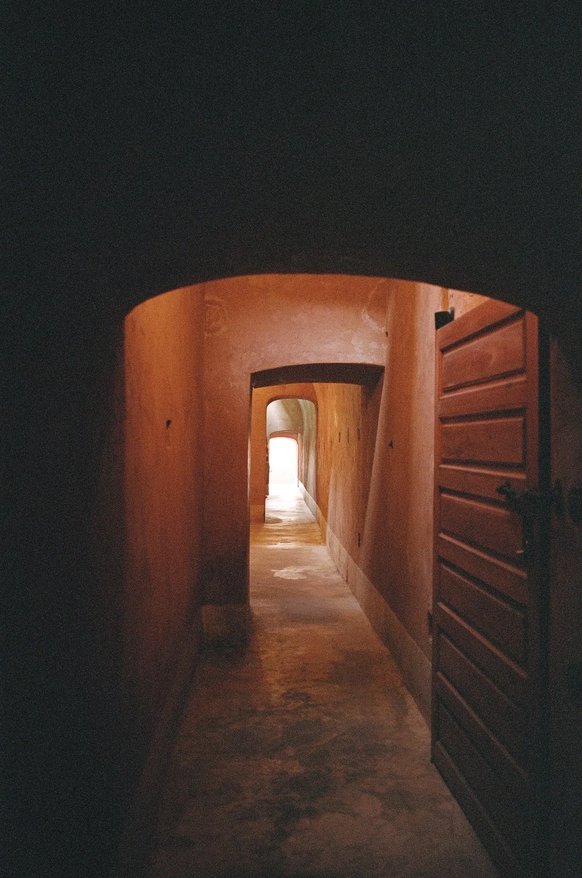 A narrow, dimly lit hallway with warm-colored walls, an arched doorway, and wooden accents, leading to a bright room at the end.