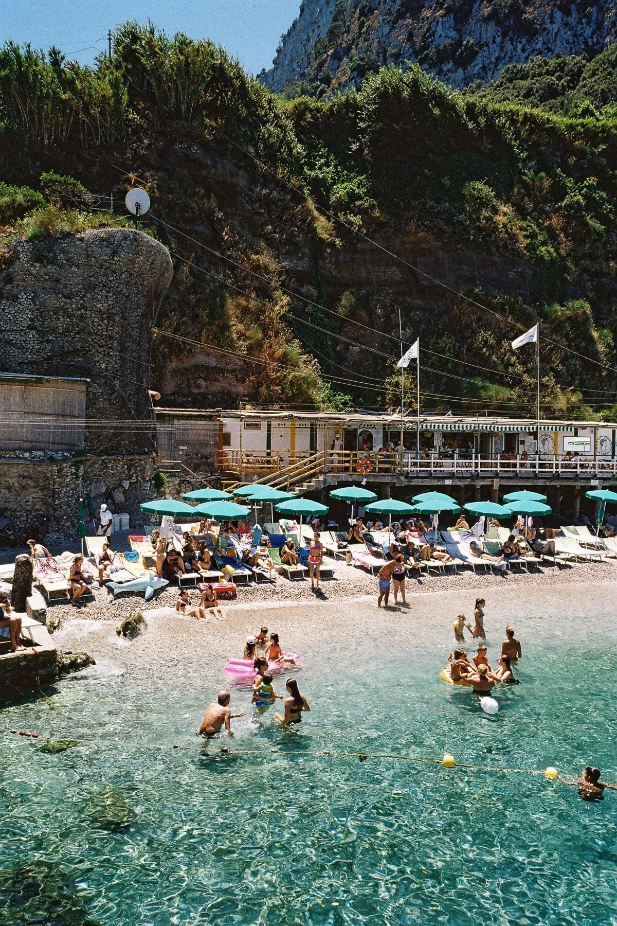 People enjoying a beach with umbrellas and lounge chairs, swimming, and playing in the water against a backdrop of rocky cliffs and lush greenery.
