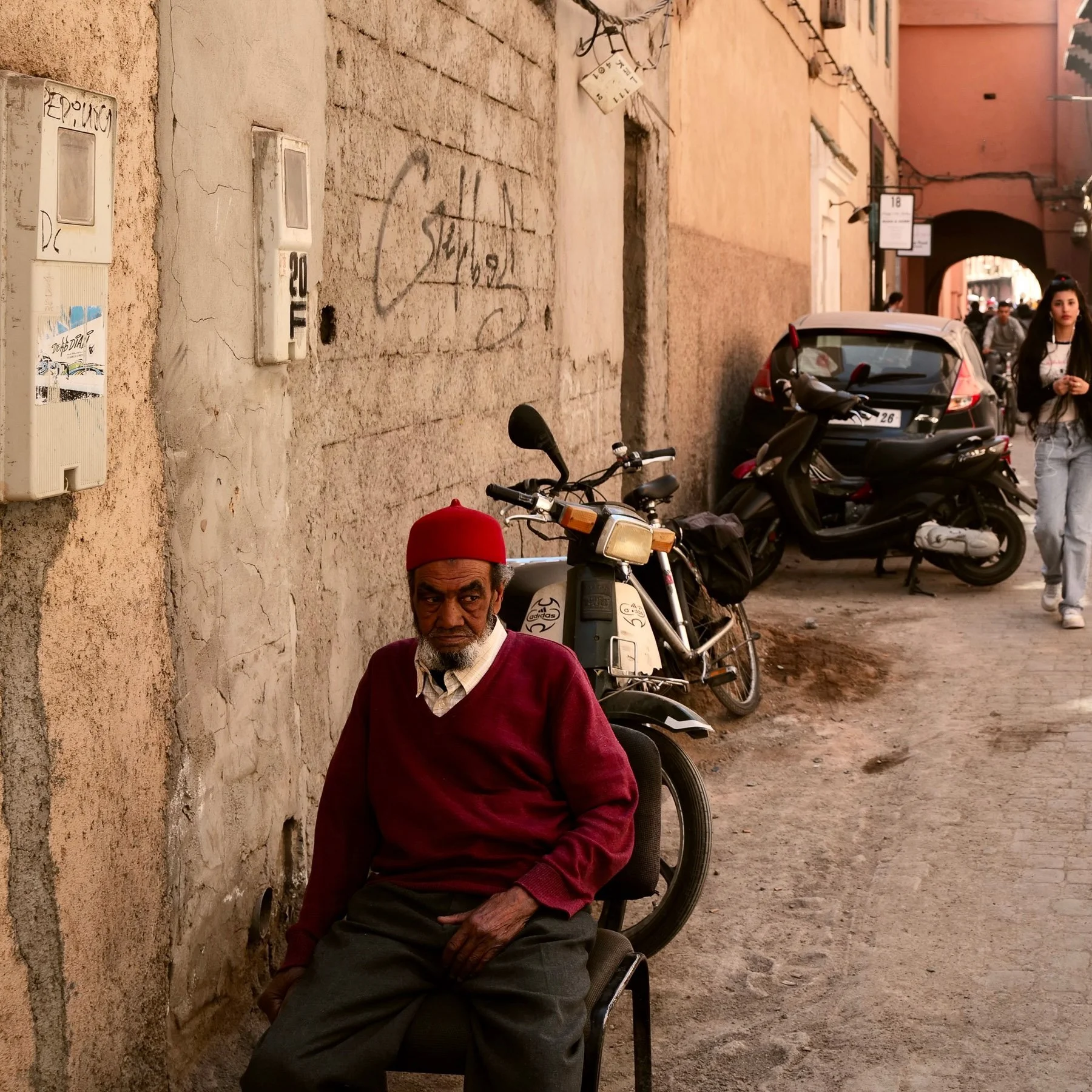 An elderly man wearing a red hat and maroon sweater sitting on a chair next to a wall. Several motorcycles and a car are parked nearby. A woman walking with earphones in her ears can be seen in the background.