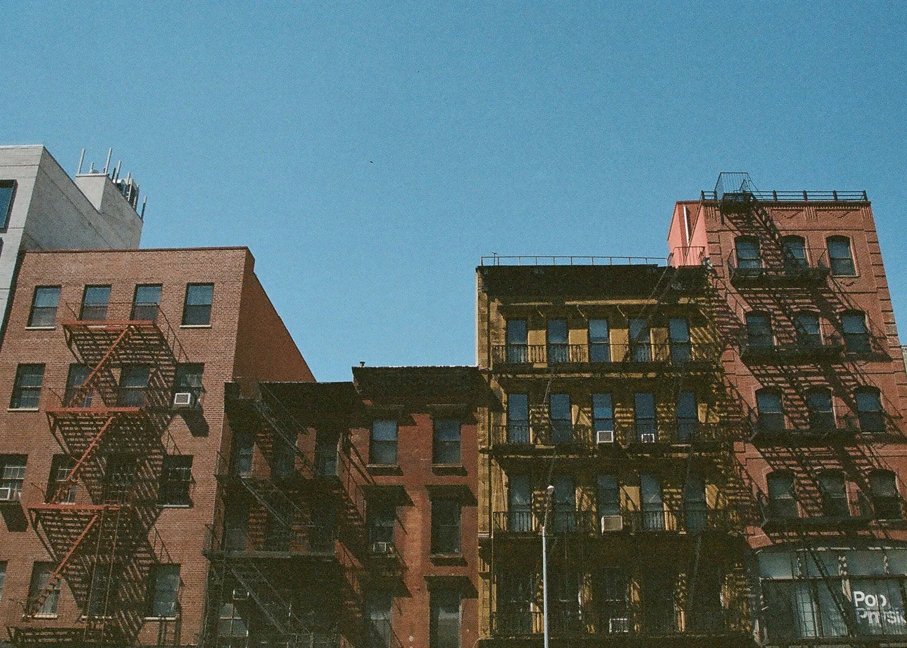 Several multistory brick buildings with fire escapes on the exterior, under a clear blue sky.
