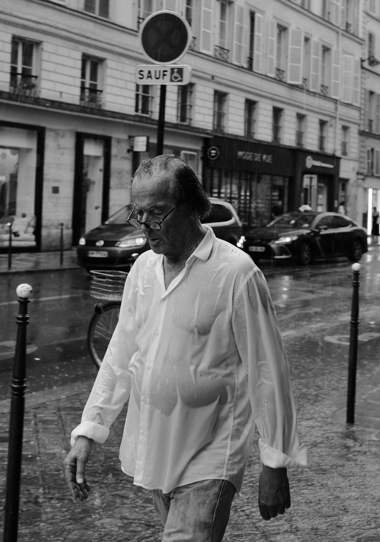 A man with glasses and wet hair walking on a city street in rain, wearing a white shirt that is also wet.