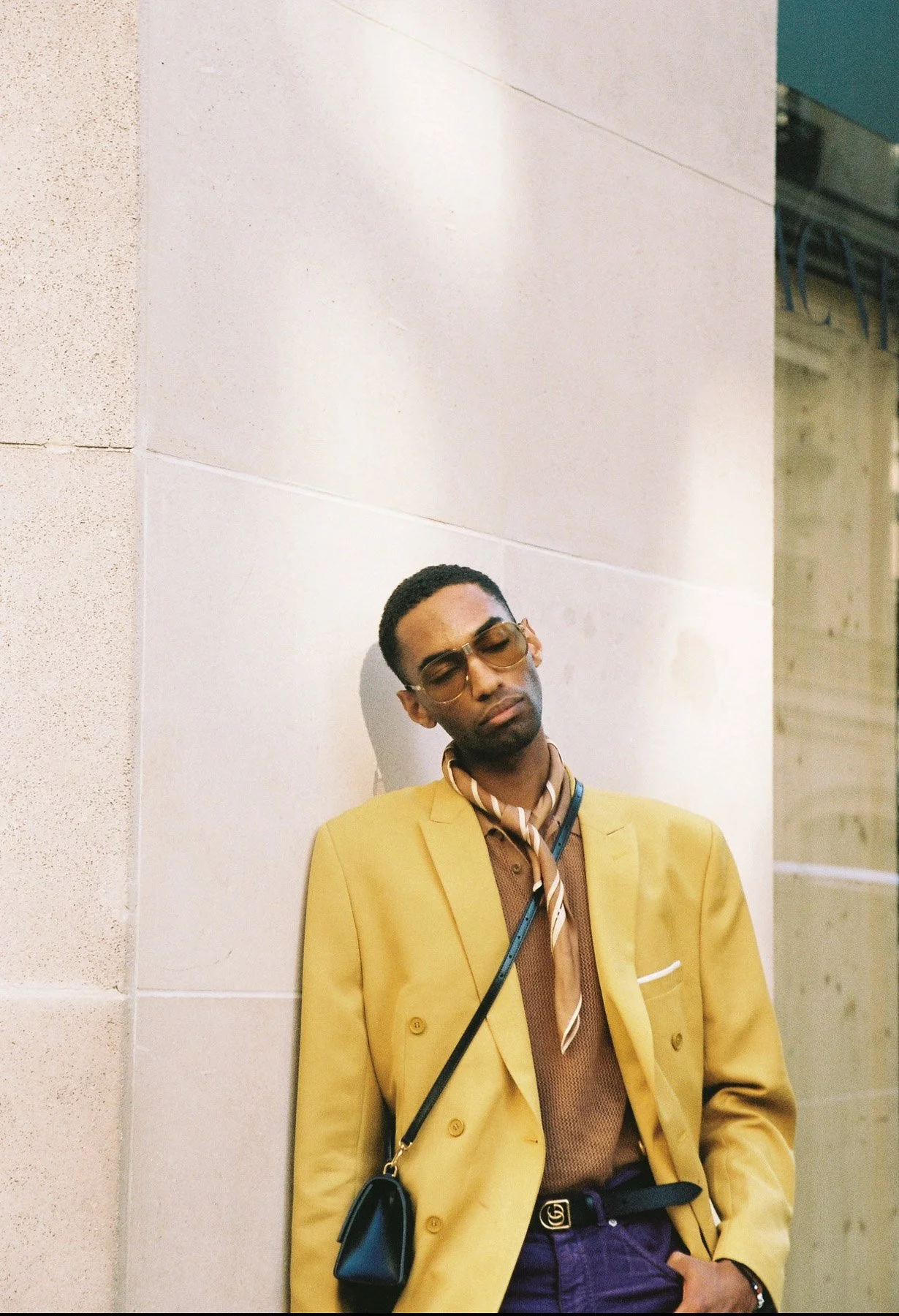 A young man with dark skin, short black hair, and wearing sunglasses, a mustard yellow blazer, a brown shirt, a beige and white striped scarf, and a black crossbody bag. He is standing against a white wall, with his eyes closed and head tilted slight