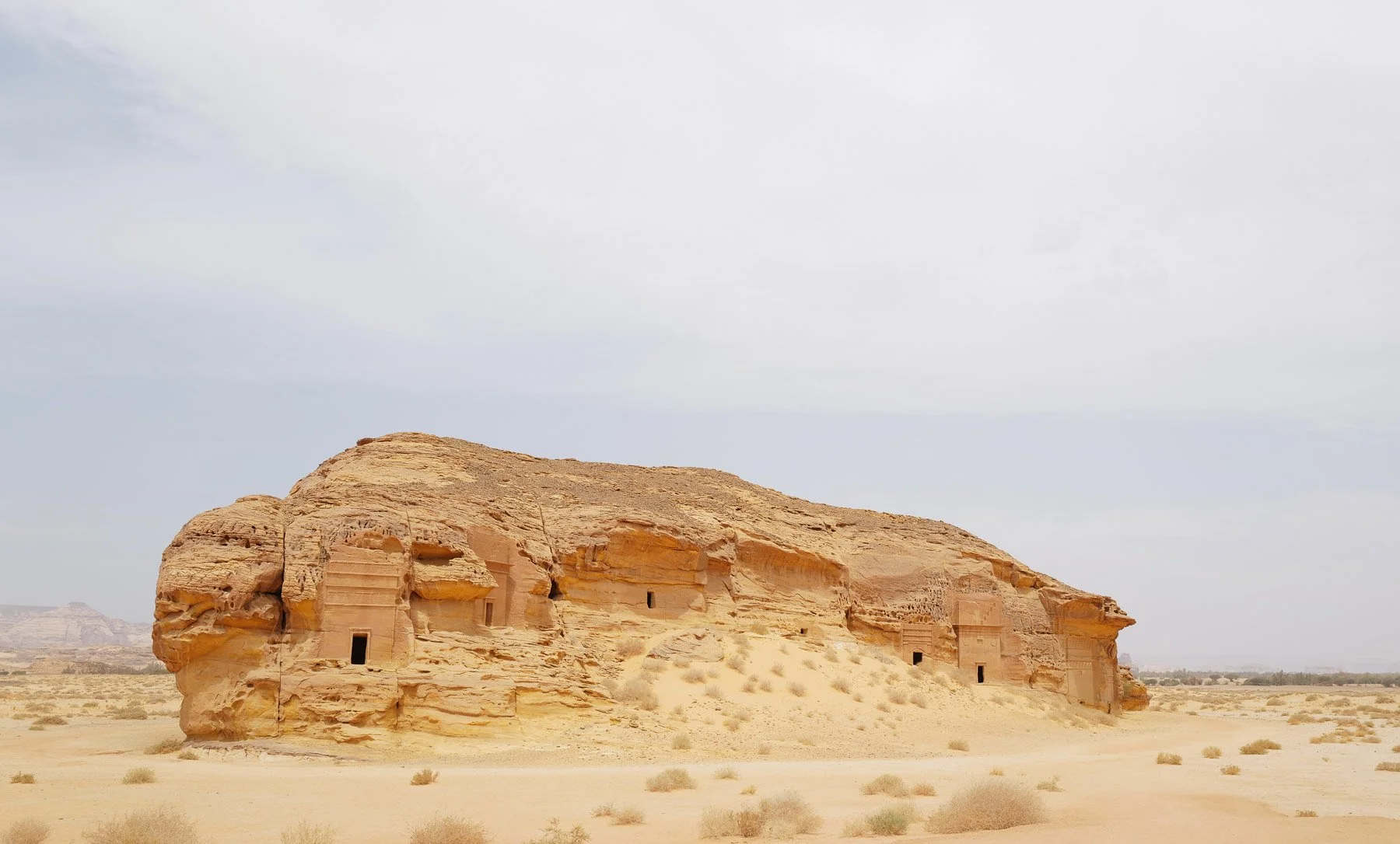 Ancient desert stone structures carved into a large sandstone rock formation under a cloudy sky, in a remote arid landscape.