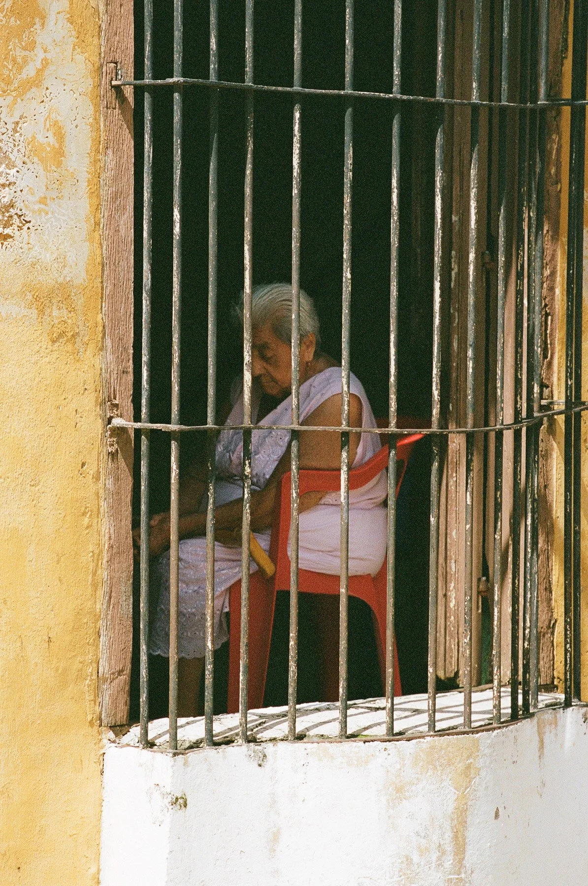 An elderly woman with silver hair sitting behind iron bars on a window, against a yellow and weathered wall, reading a book.