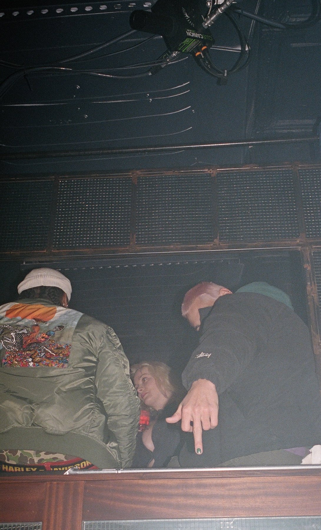 Two individuals are leaning over and engaging with a woman lying on the ground. The woman with blonde hair appears to be listening or talking to the men, who are pointing and leaning in. The setting is indoors, with visible wires, a camera, and a met
