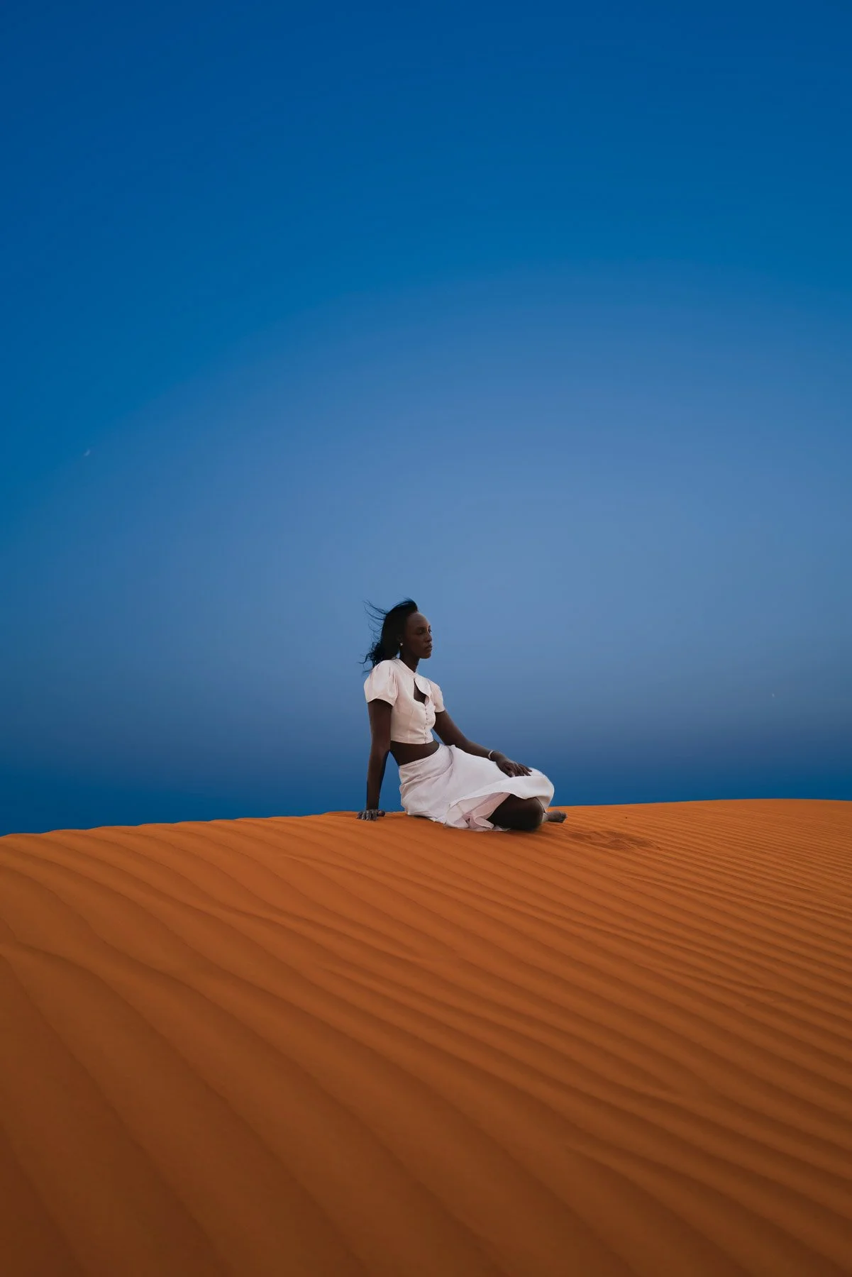 A woman sitting on orange sand dunes in a white dress, with a deep blue sky overhead.