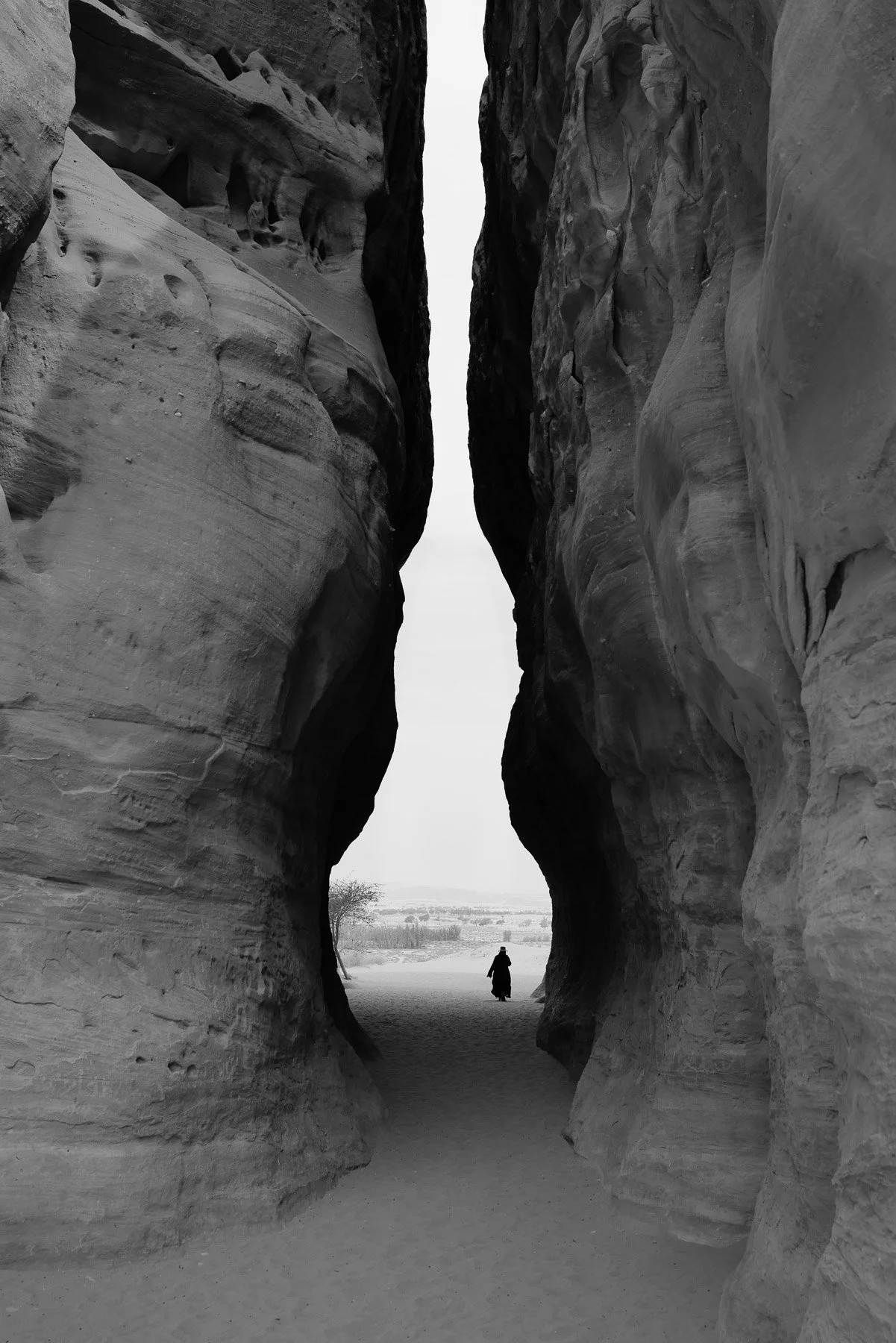 Black and white photo of a narrow canyon with a person walking through it, viewed from within the canyon, with the canyon walls towering on either side.