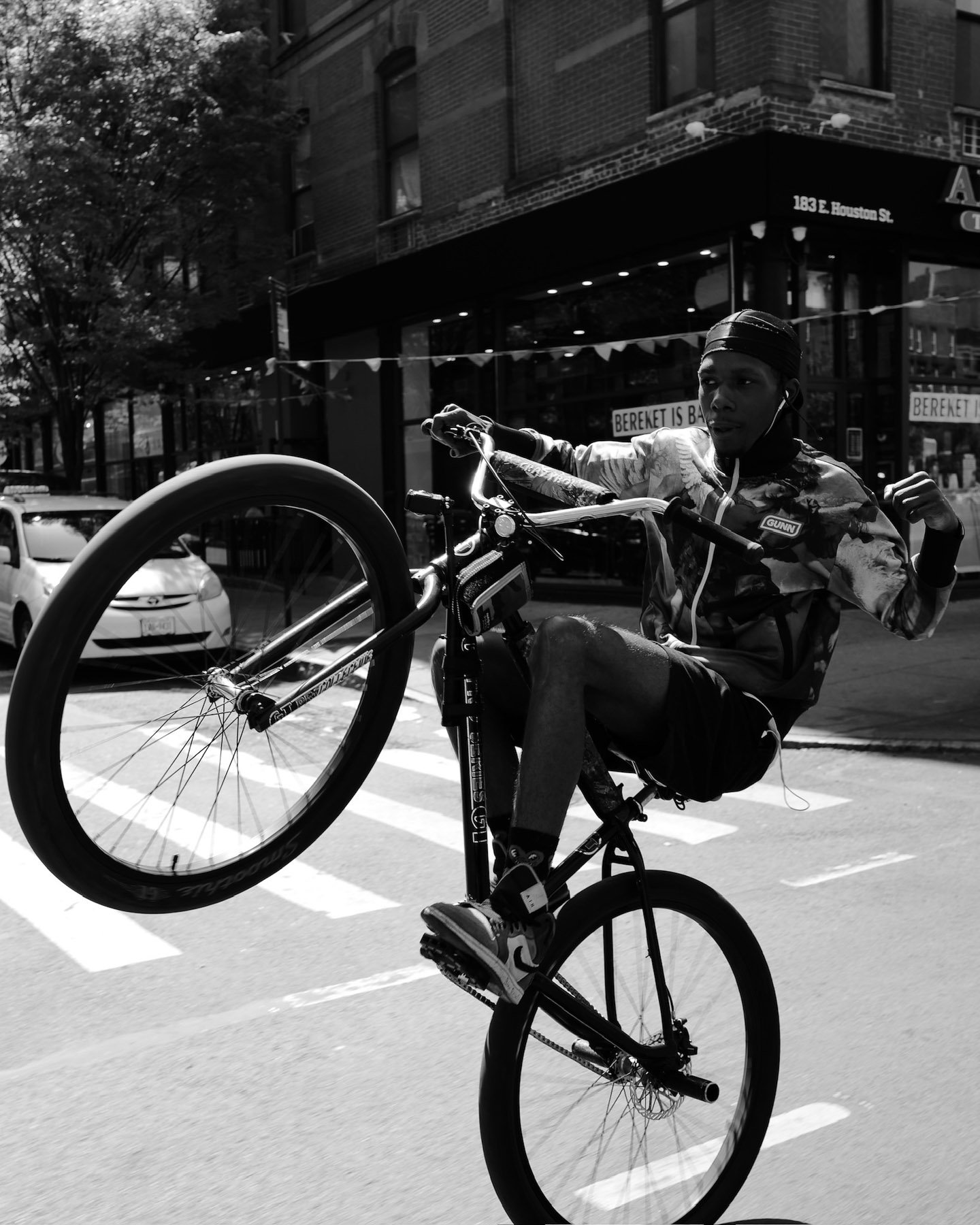 A person performing a wheelie stunt on a bicycle in an urban street, with buildings and parked cars in the background.