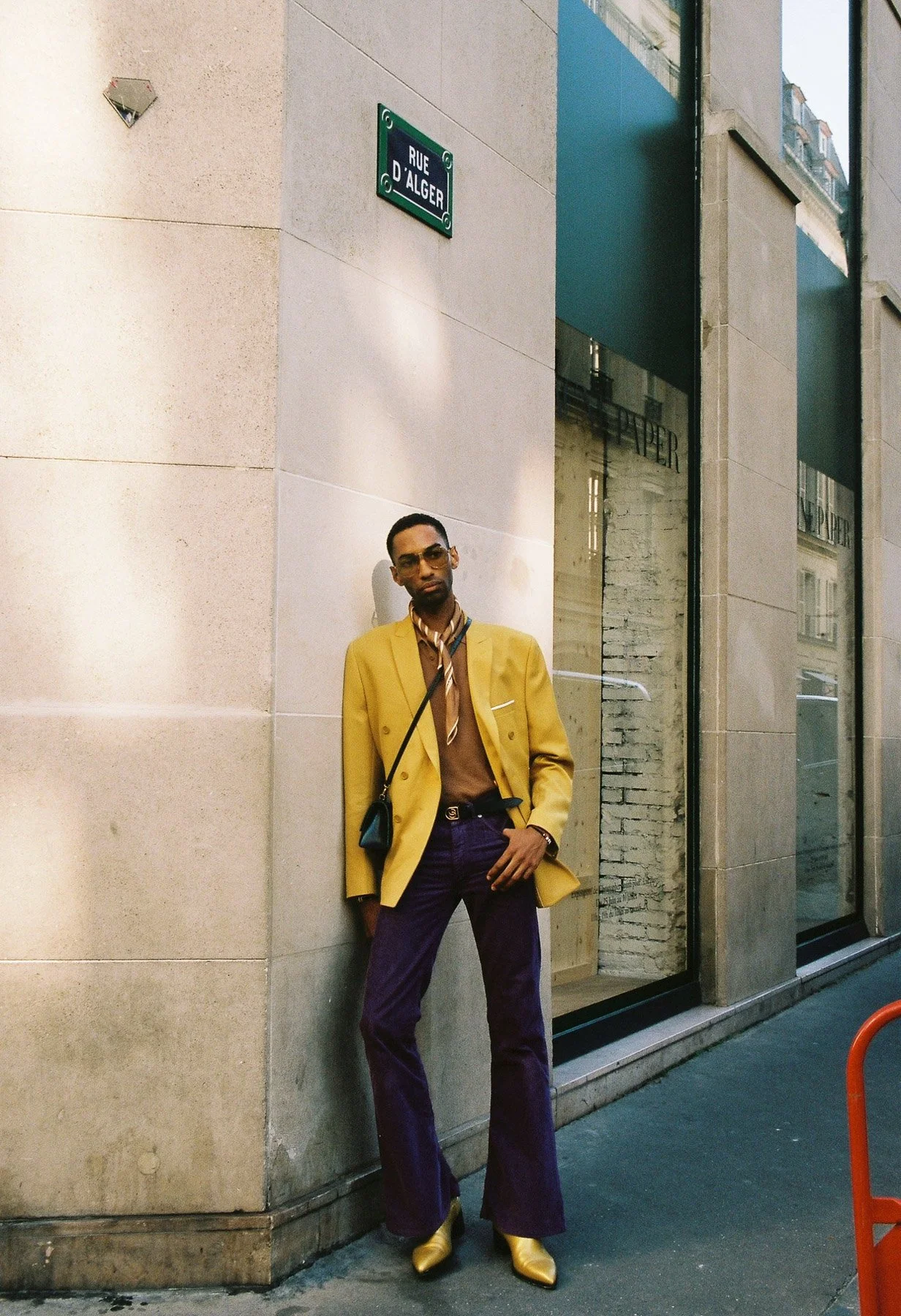 A man standing against a beige wall on a city street, wearing a yellow blazer, purple pants, gold boots, sunglasses, and a patterned scarf.