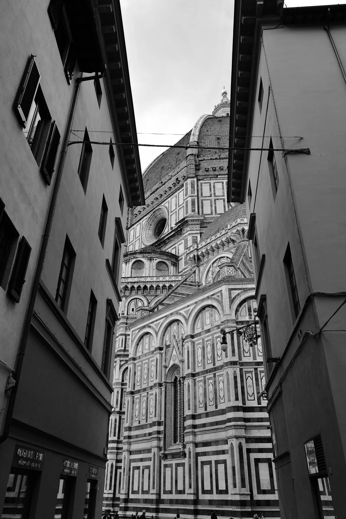 Black and white photo of the Florence Cathedral, also known as the Duomo, viewed from a narrow street framed by buildings. The cathedral's ornate facade and large dome are visible in the background.