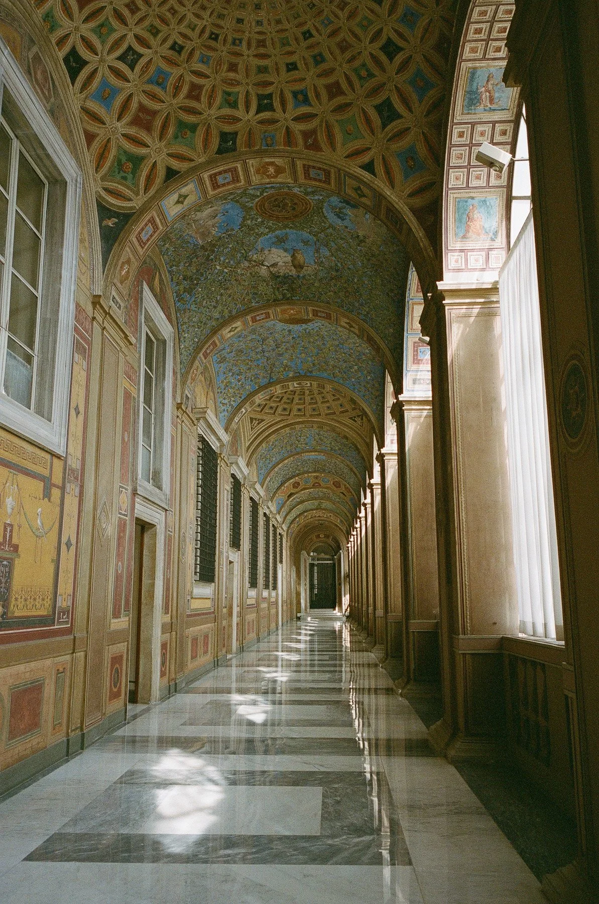 A long, ornate corridor with decorated vaulted ceilings, tall windows, and marble flooring, in a historic or elegant building.