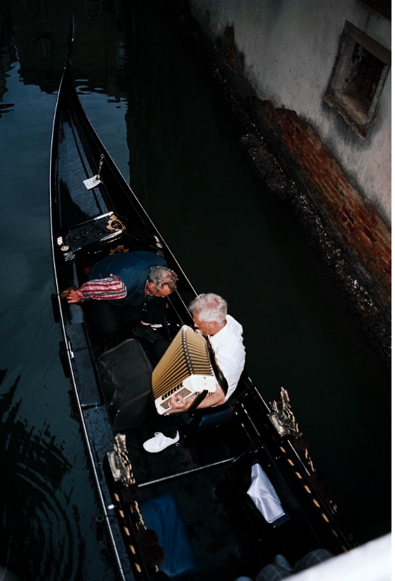 Two men in a gondola, one holding an accordion, floating in a narrow canal with a brick wall and a small window along the side.