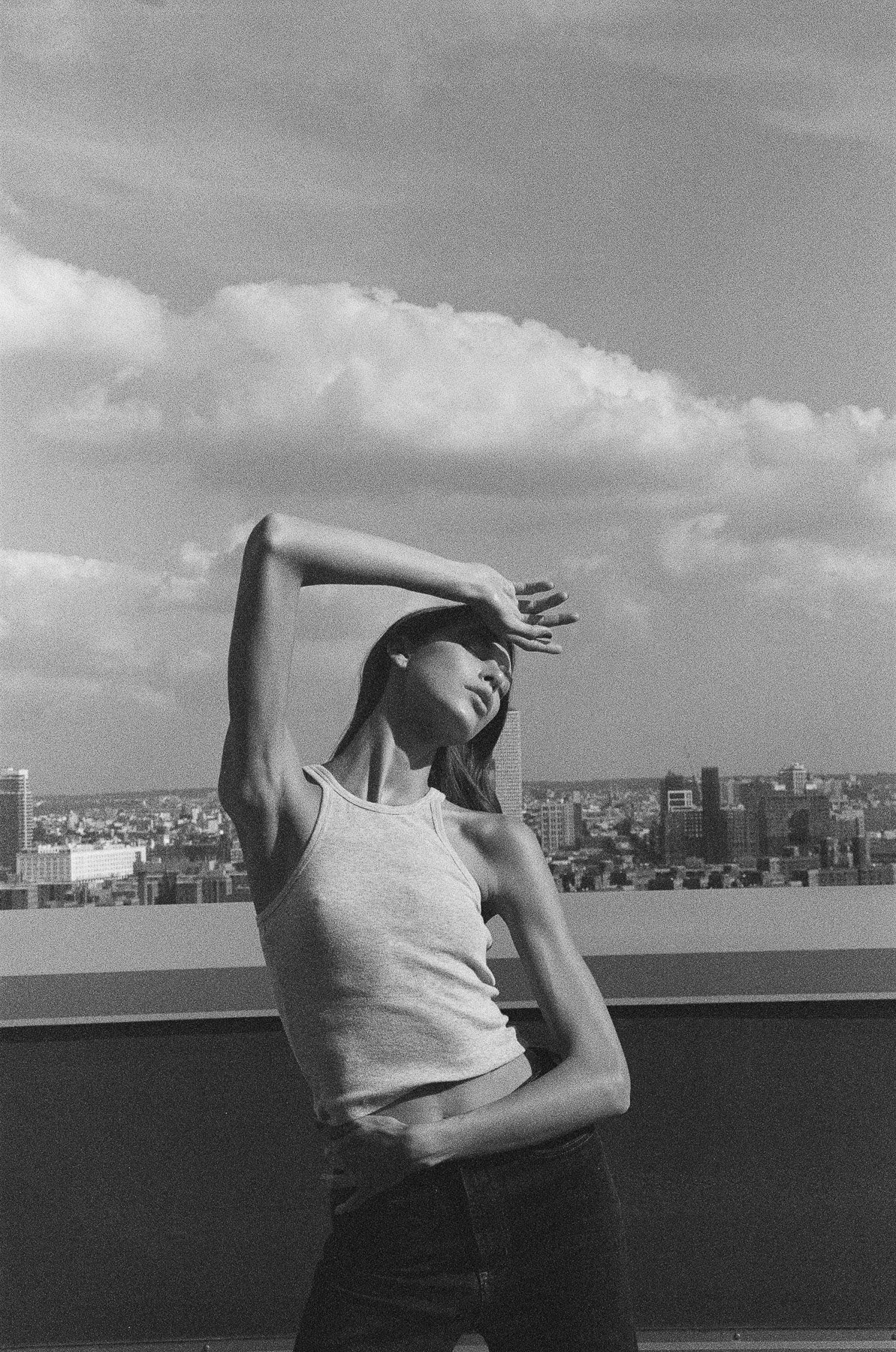 A woman with long hair wearing a sleeveless top and dark pants, posing outdoors on a rooftop with a city skyline in the background, shielding her eyes from the sun with her right hand.