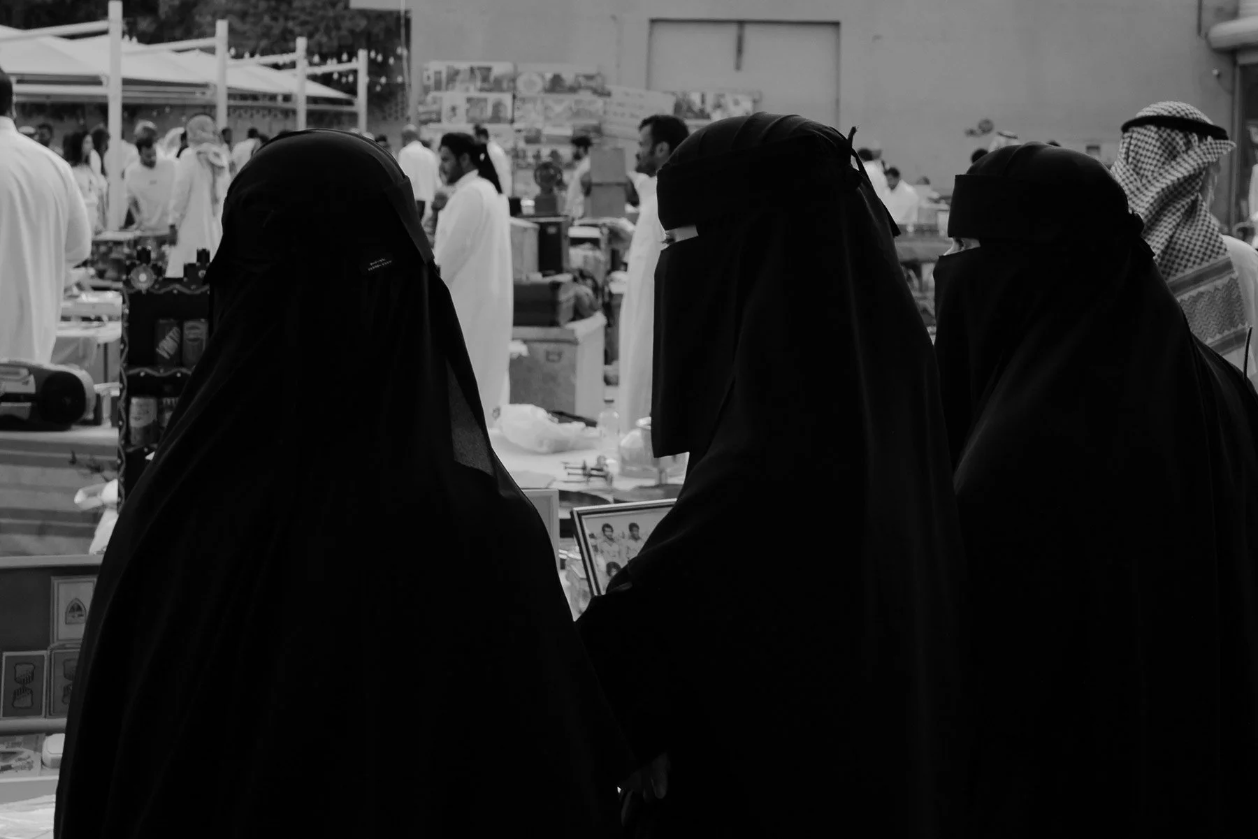 Three women wearing black abayas at an outdoor market, interacting with a display or photo, with people and market stalls in the background.