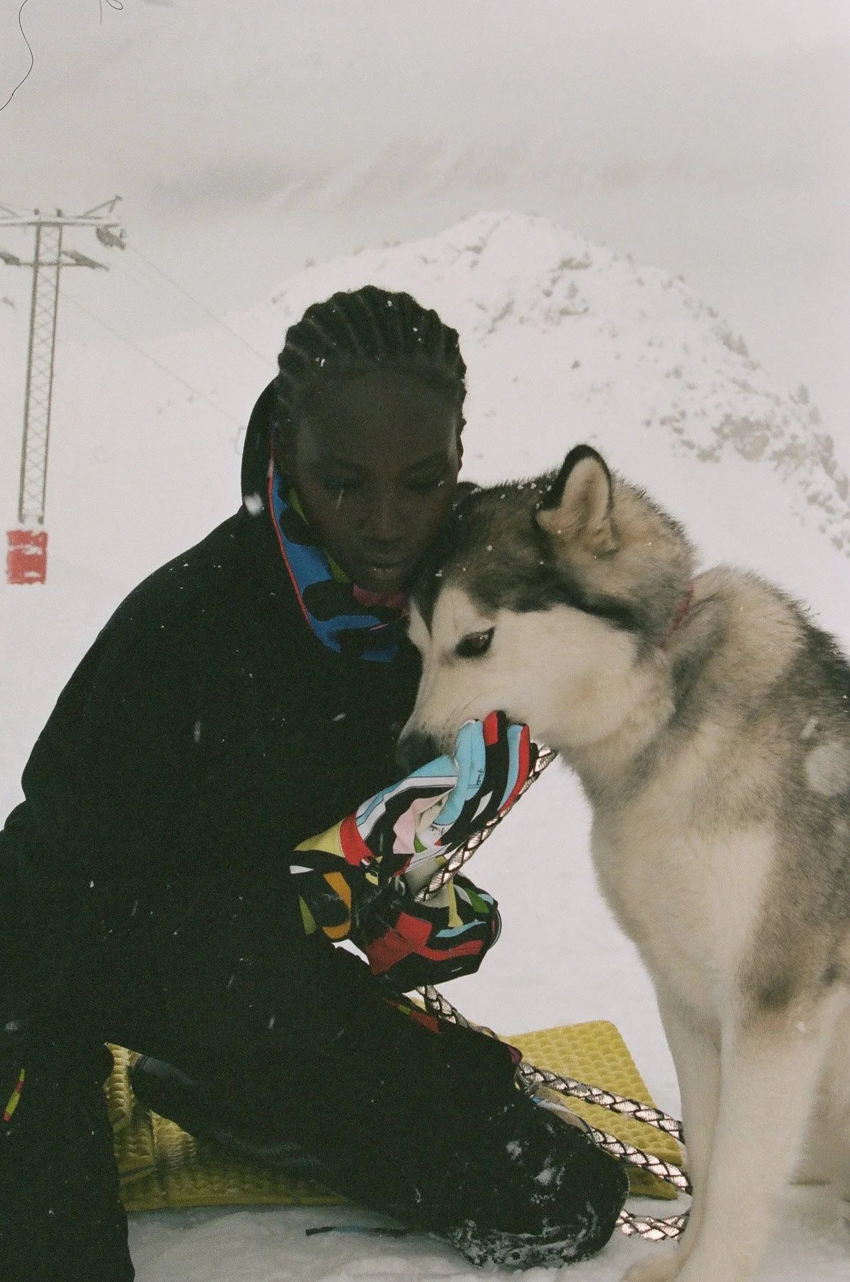 A person with dark skin, dressed in winter clothing, including colorful snow gloves, kneeling in the snow and saluting a Siberian Husky dog. The background features snowy mountains and a ski lift in a winter landscape.