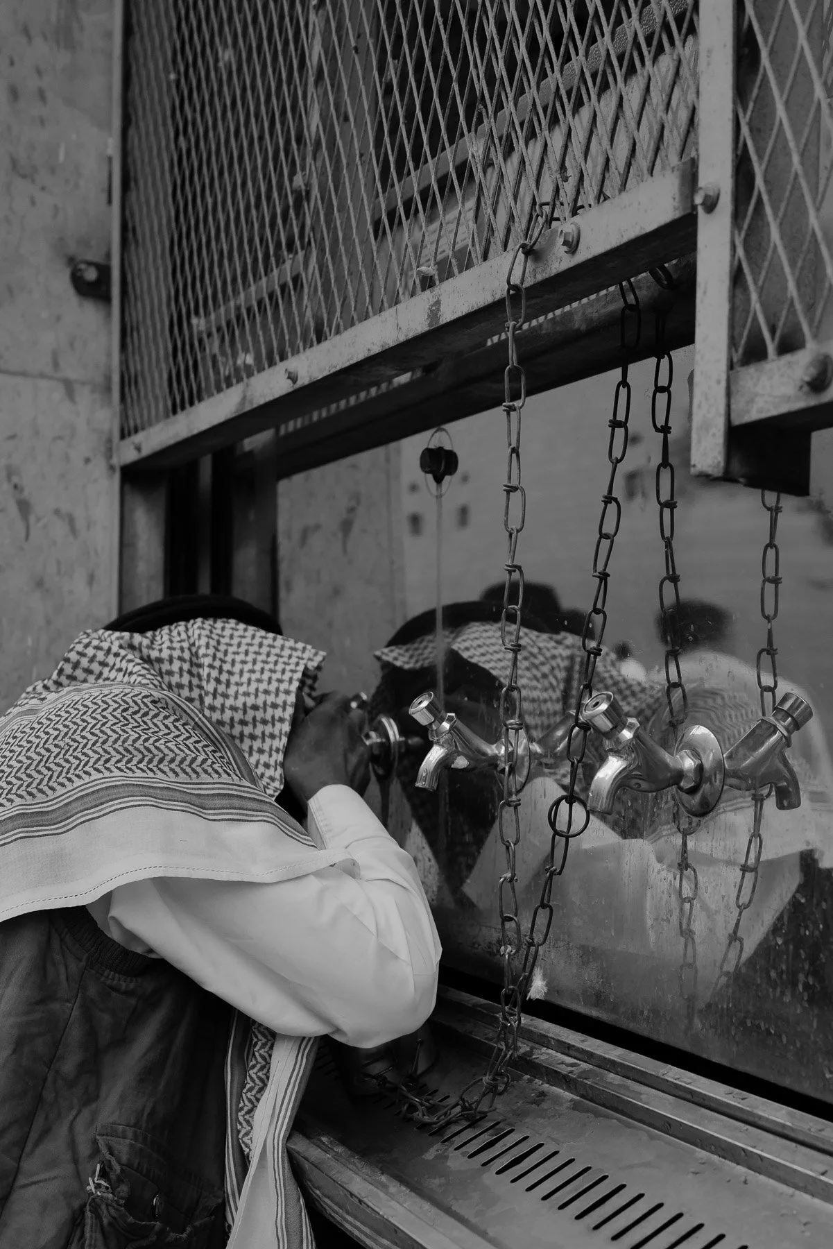 A person wearing a traditional Middle Eastern headscarf is pouring water from a tap on a street sidewalk into a container near a fenced building.
