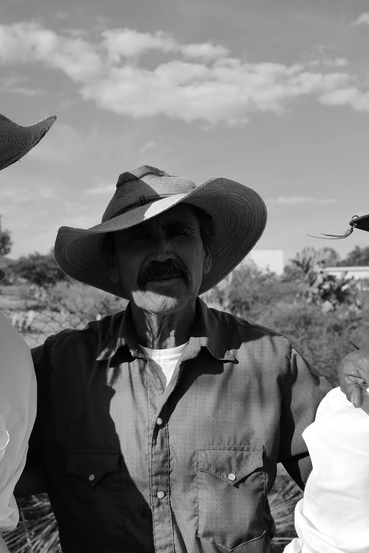 A man wearing a wide-brimmed straw hat and a button-up shirt, standing outdoors with a background of trees and a partly cloudy sky.