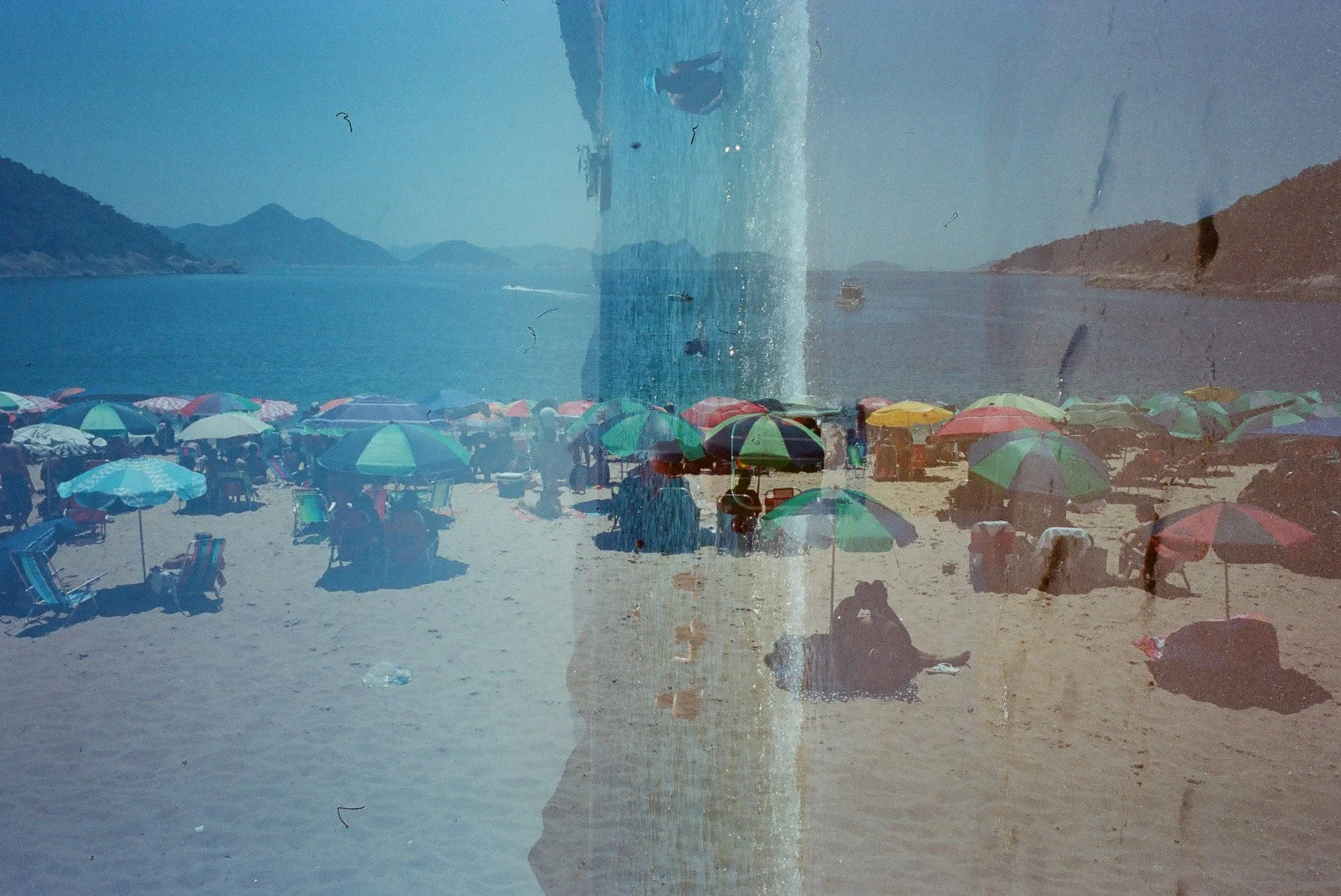 Beach scene with numerous colorful umbrellas on sandy shore, people sitting and walking, calm water with boats, and mountains in the background, viewed through a glass window with water streaks and dirt spots.