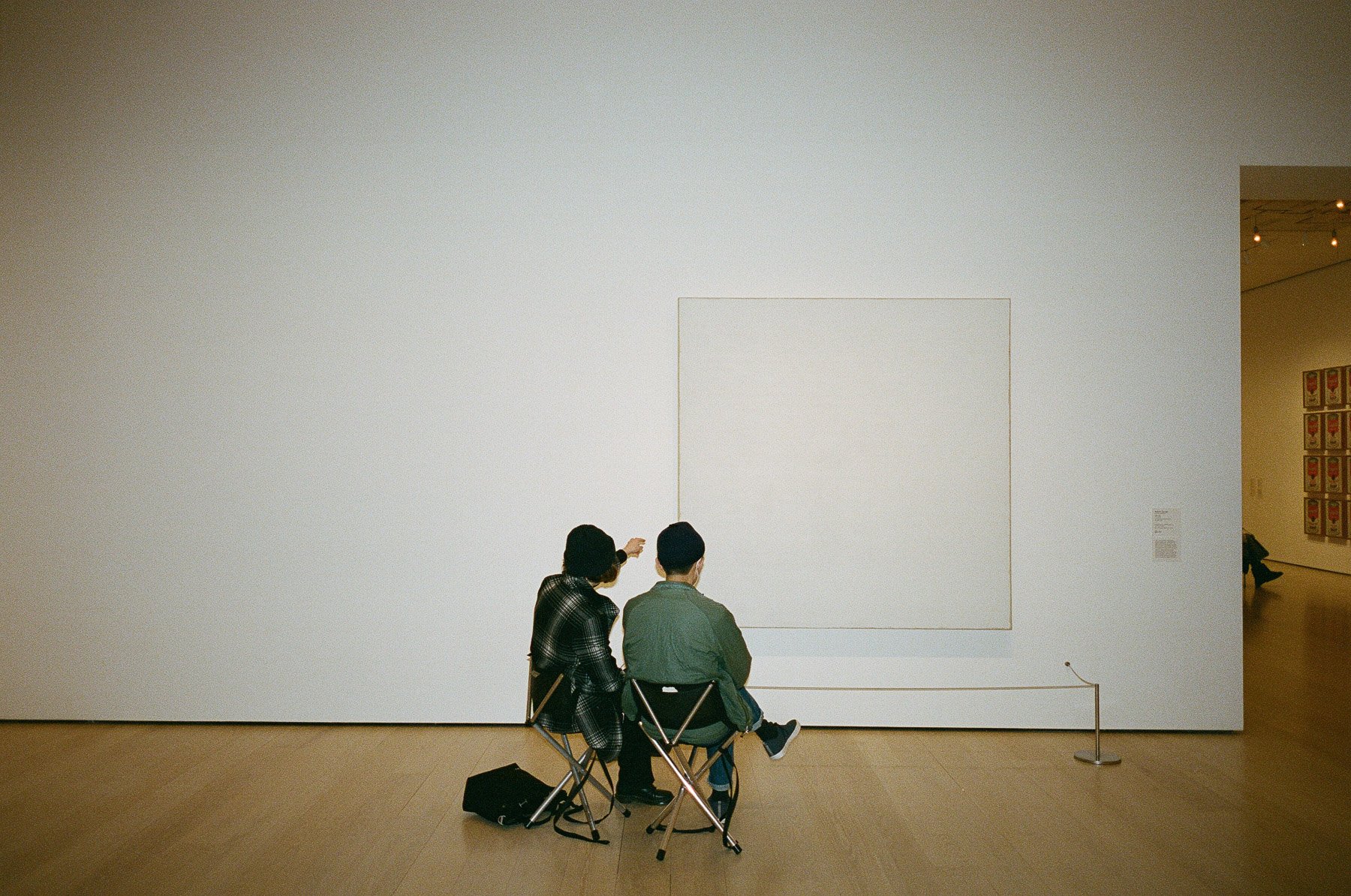 Two people sitting on chairs in an art gallery, looking at a blank square white frame on the wall. One person is pointing at the frame, and there are other artwork and gallery visitors visible in the background.